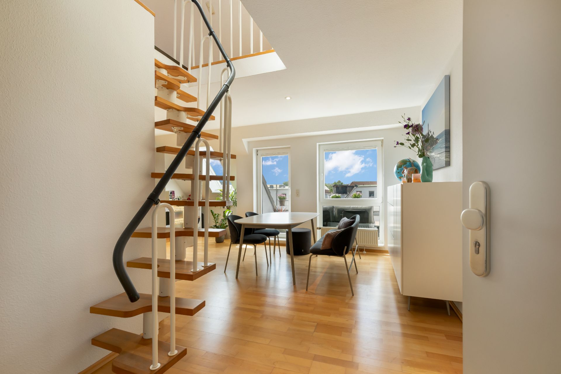 Helles Wohnzimmer mit Holztreppe, Esstisch, Stühlen und großen Fenstern.