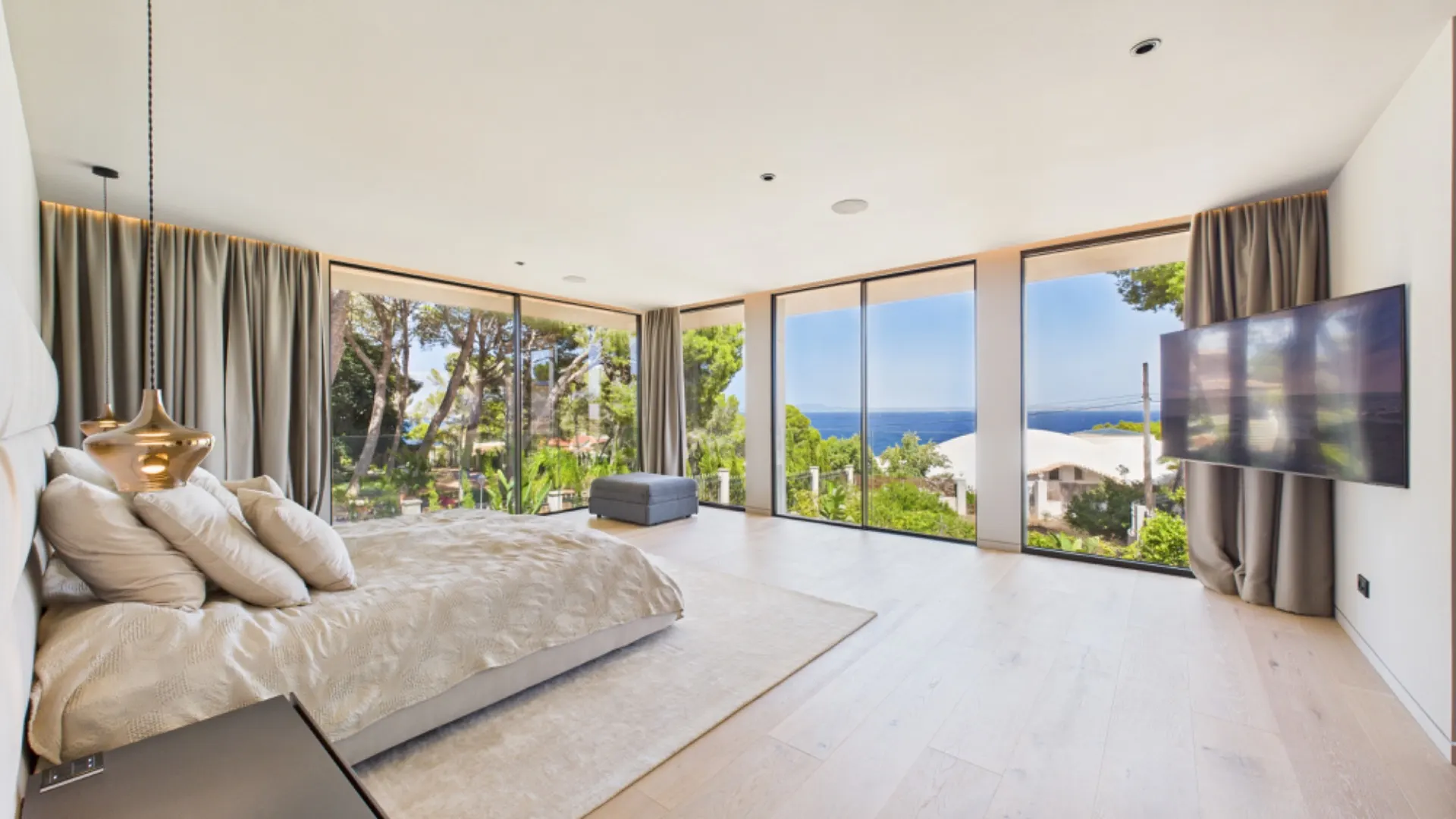 Modernes Schlafzimmer mit großem Bett, Fernseher und Panoramablick durch große Fenster auf Meer und Bäume.