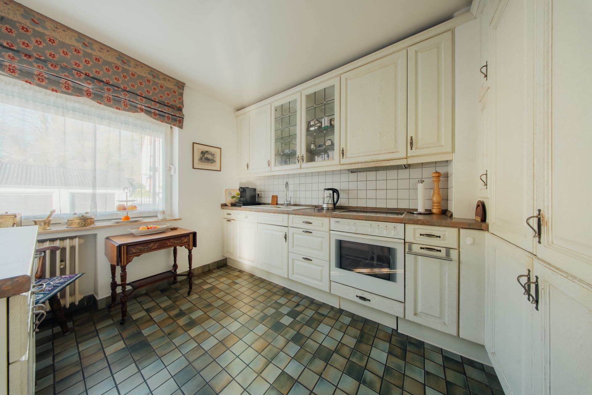 Helle Küche mit weißen Schränken, Fenster, Holztisch und gefliestem Boden.