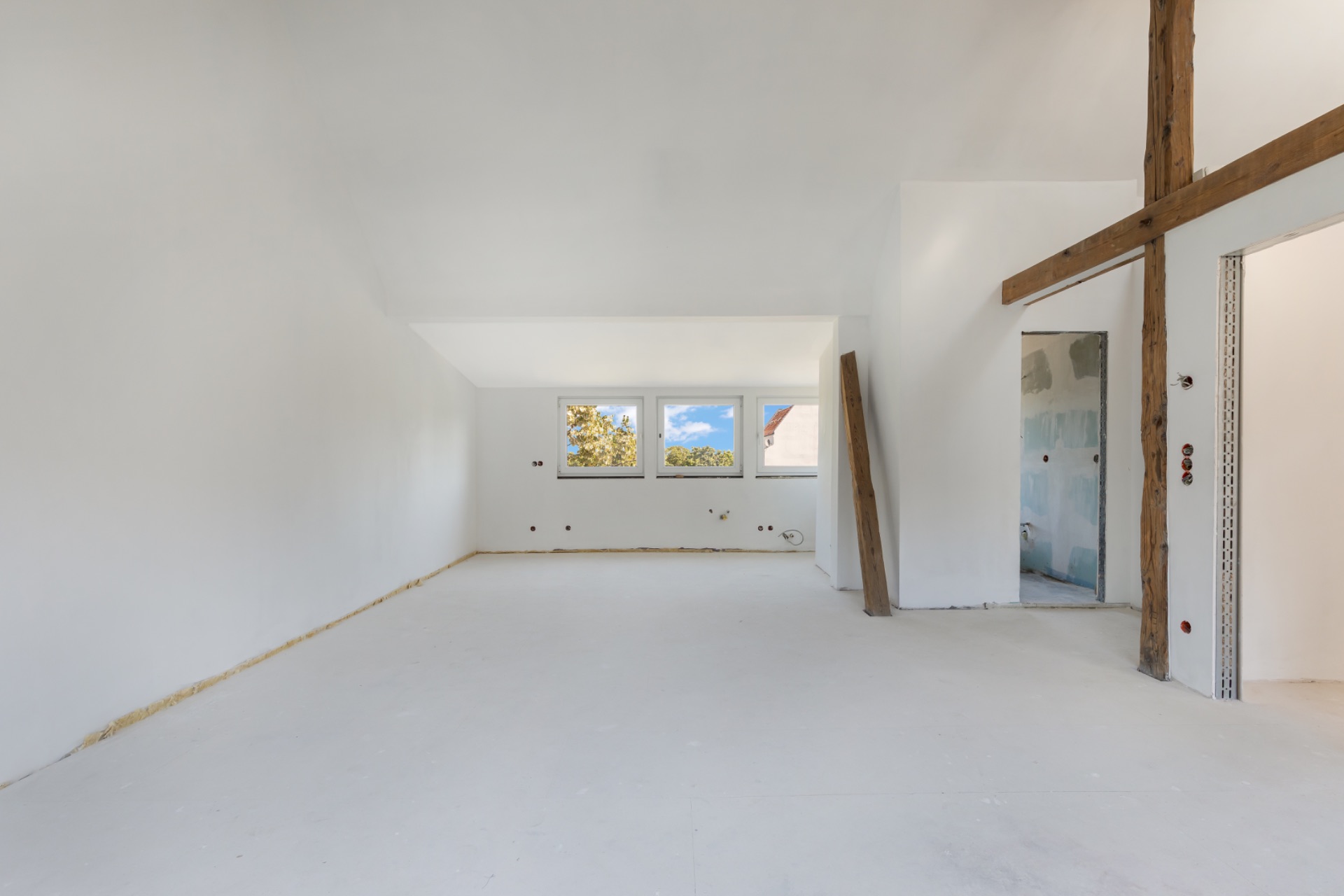Heller leerer Raum mit drei Fenstern und sichtbarem Holzbalken an der Wand.