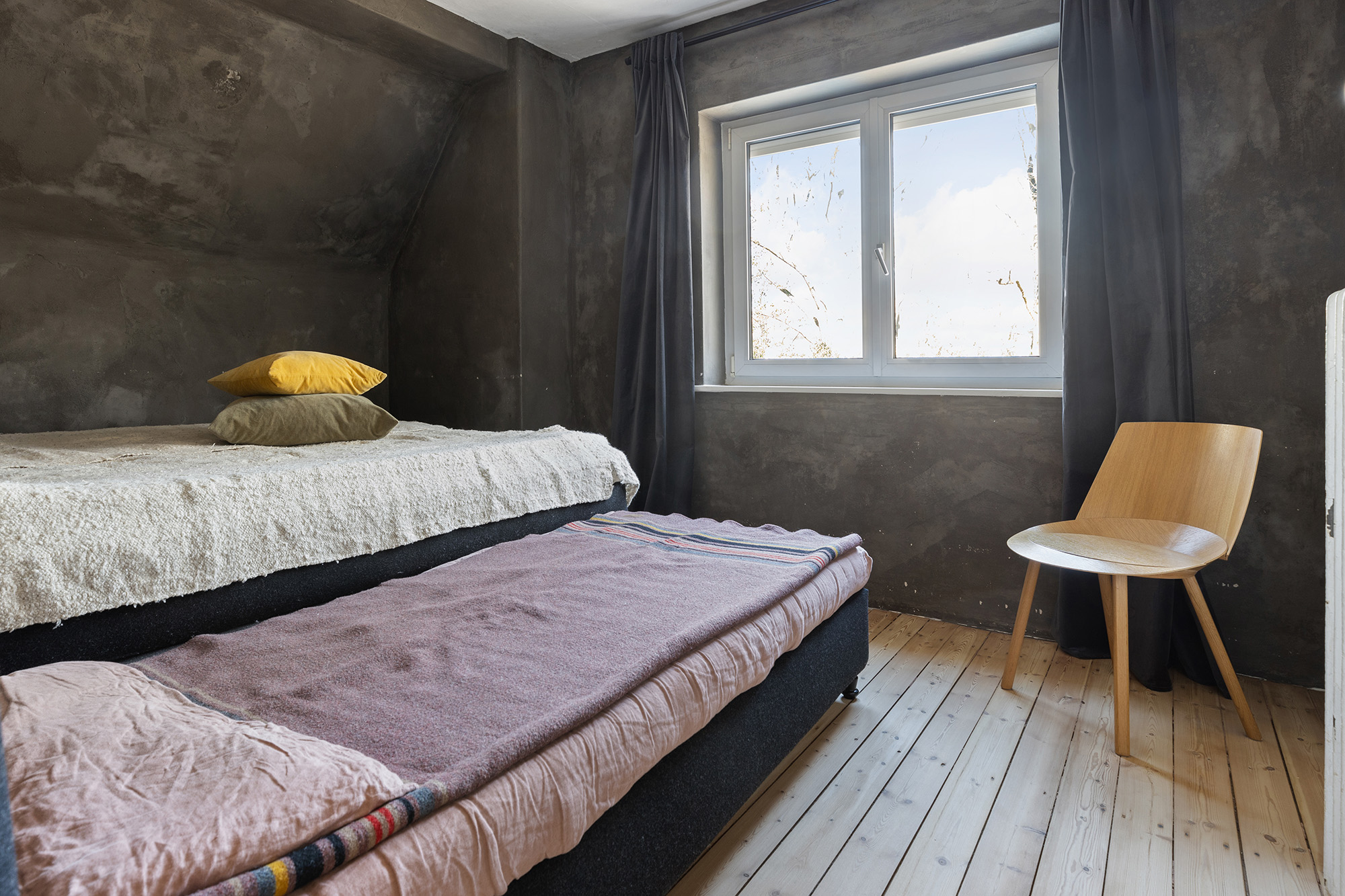 Zimmer mit zwei Einzelbetten, einem Holzstuhl und einem Fenster mit dunklen Vorhängen.