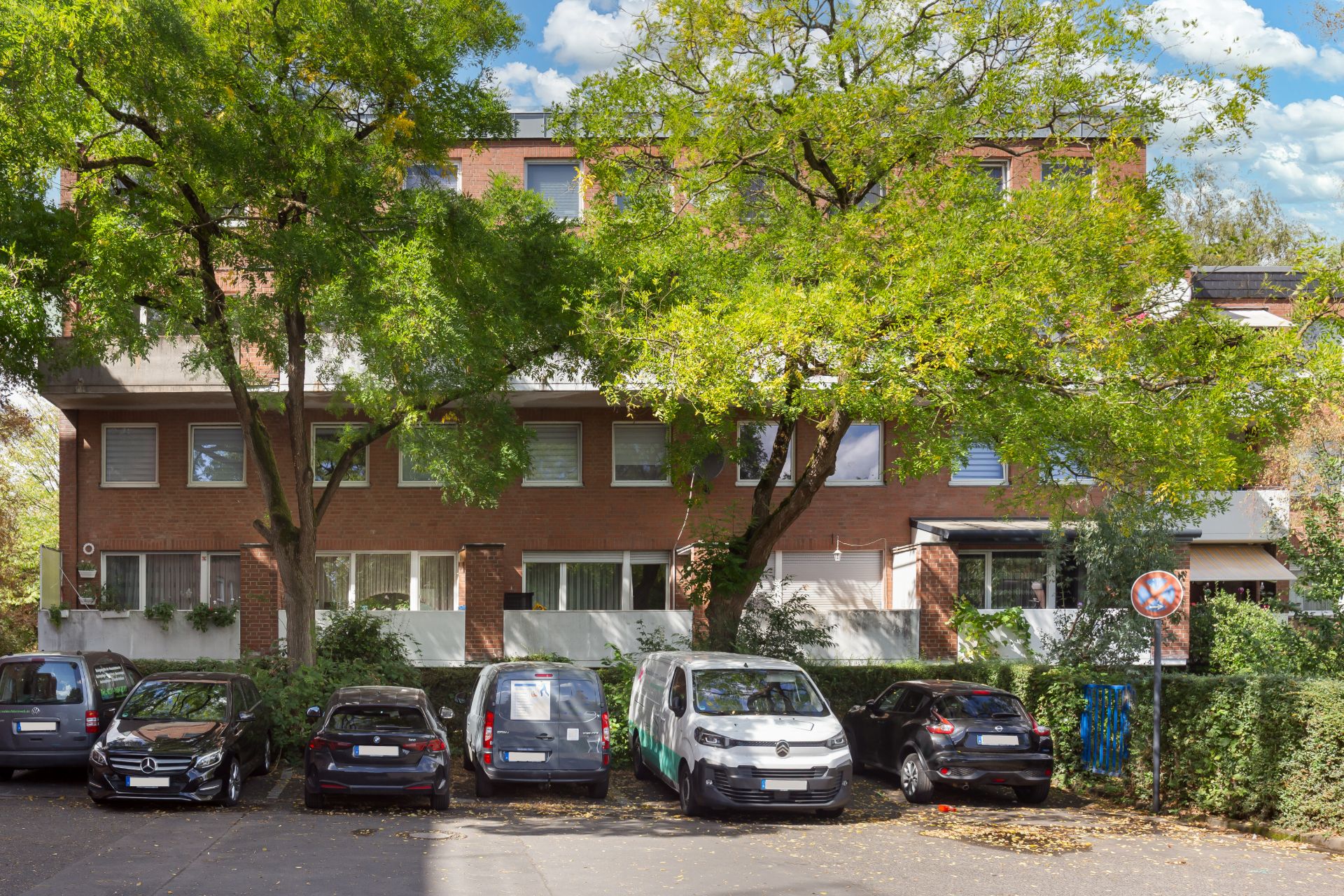 Mehrfamilienhaus mit Backsteinfassade, Bäumen und geparkten Autos davor.