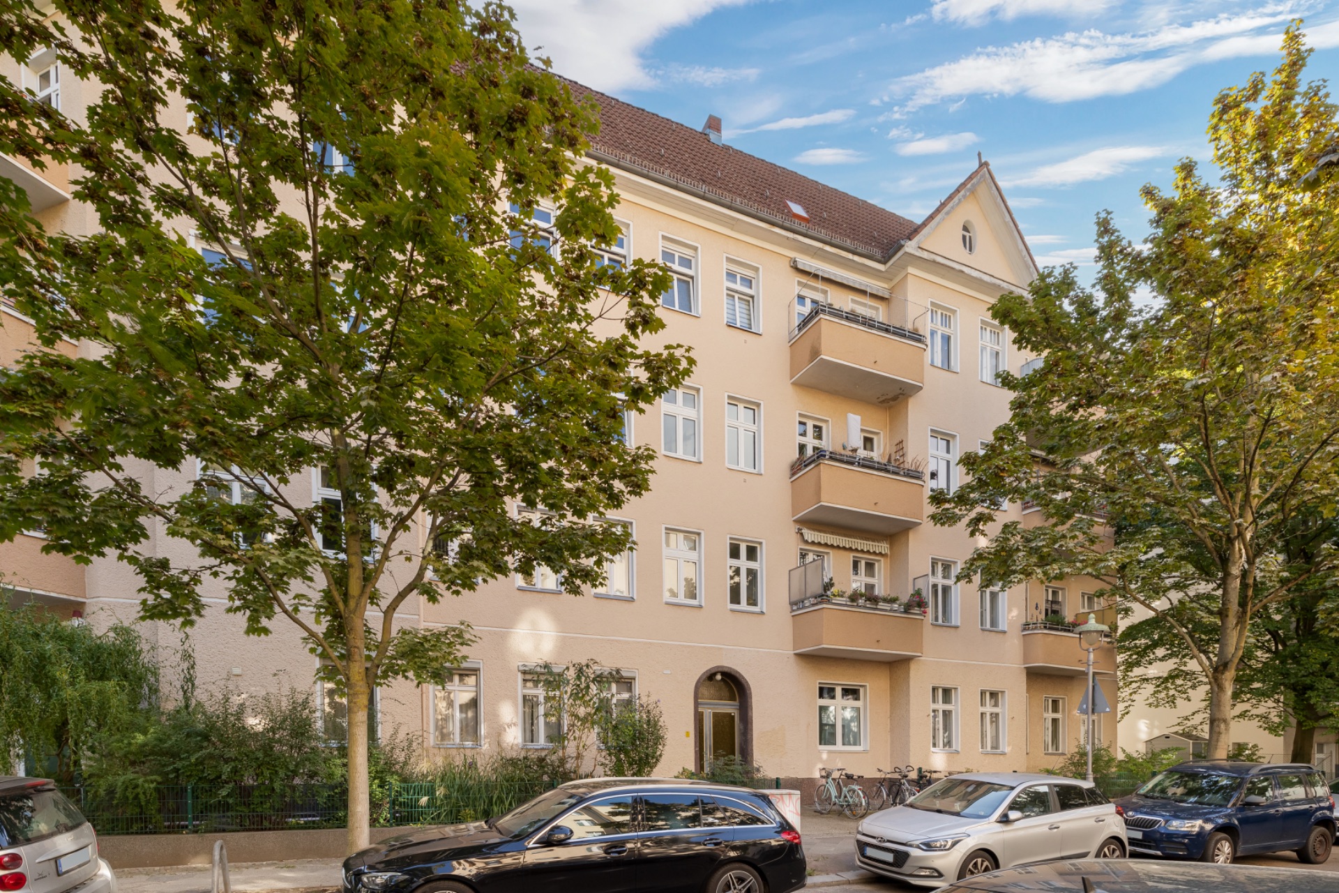Mehrfamilienhaus mit Balkonen, Bäumen davor und geparkten Autos an der Straße.