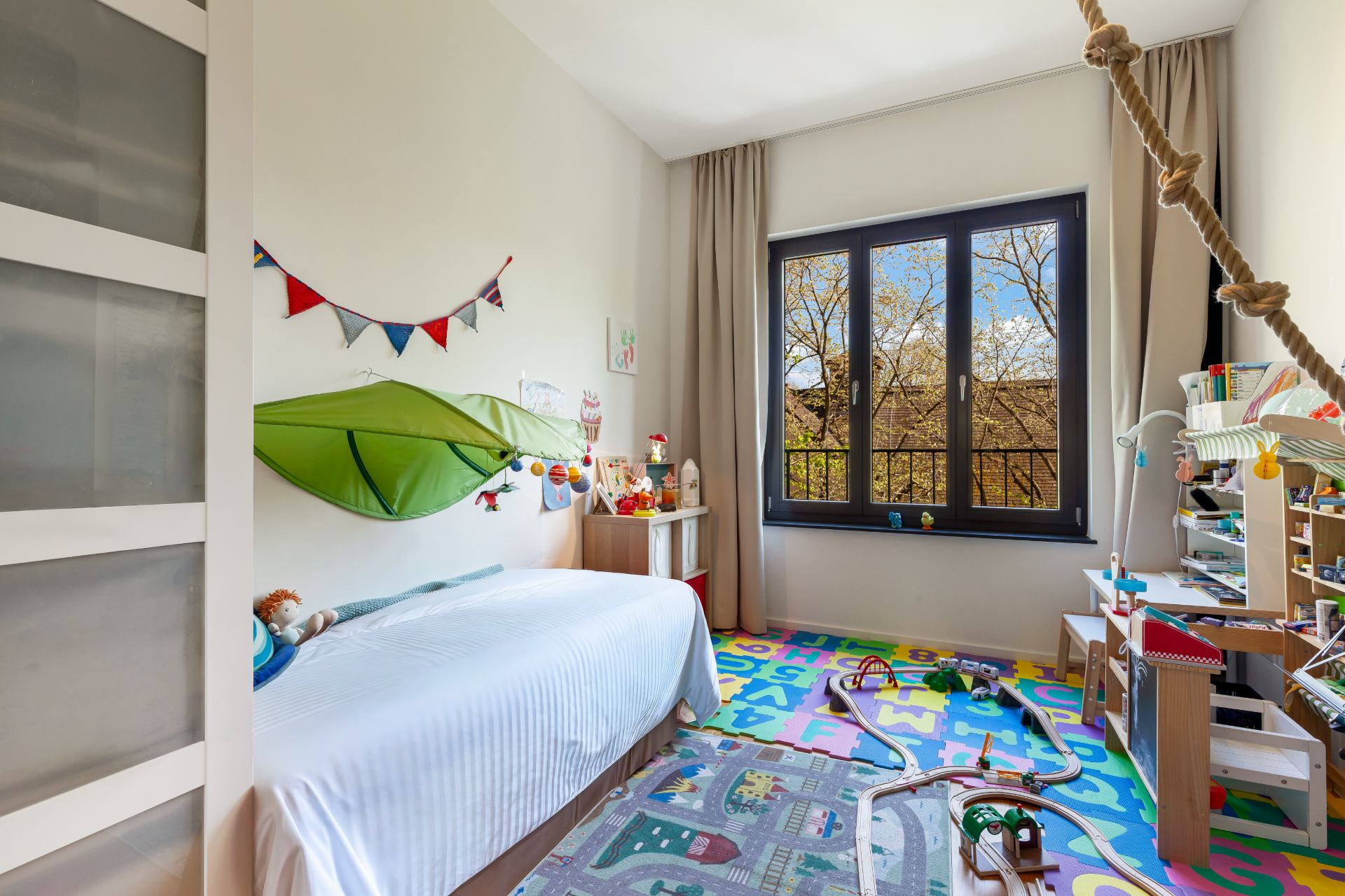 Helles Kinderzimmer mit Spielzeug, Teppich, Hochbett und großem Fenster.