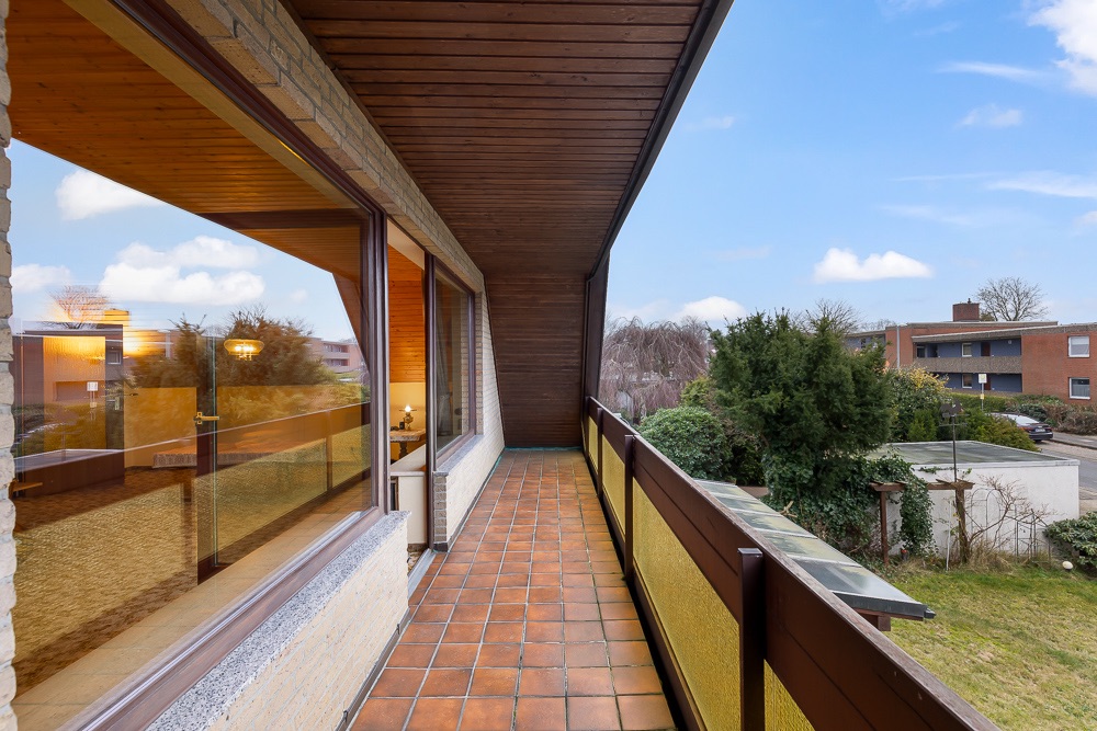 Langer Balkon mit braunen Fliesen, Holzdach und Blick auf Nachbargärten und Wohnhäuser.