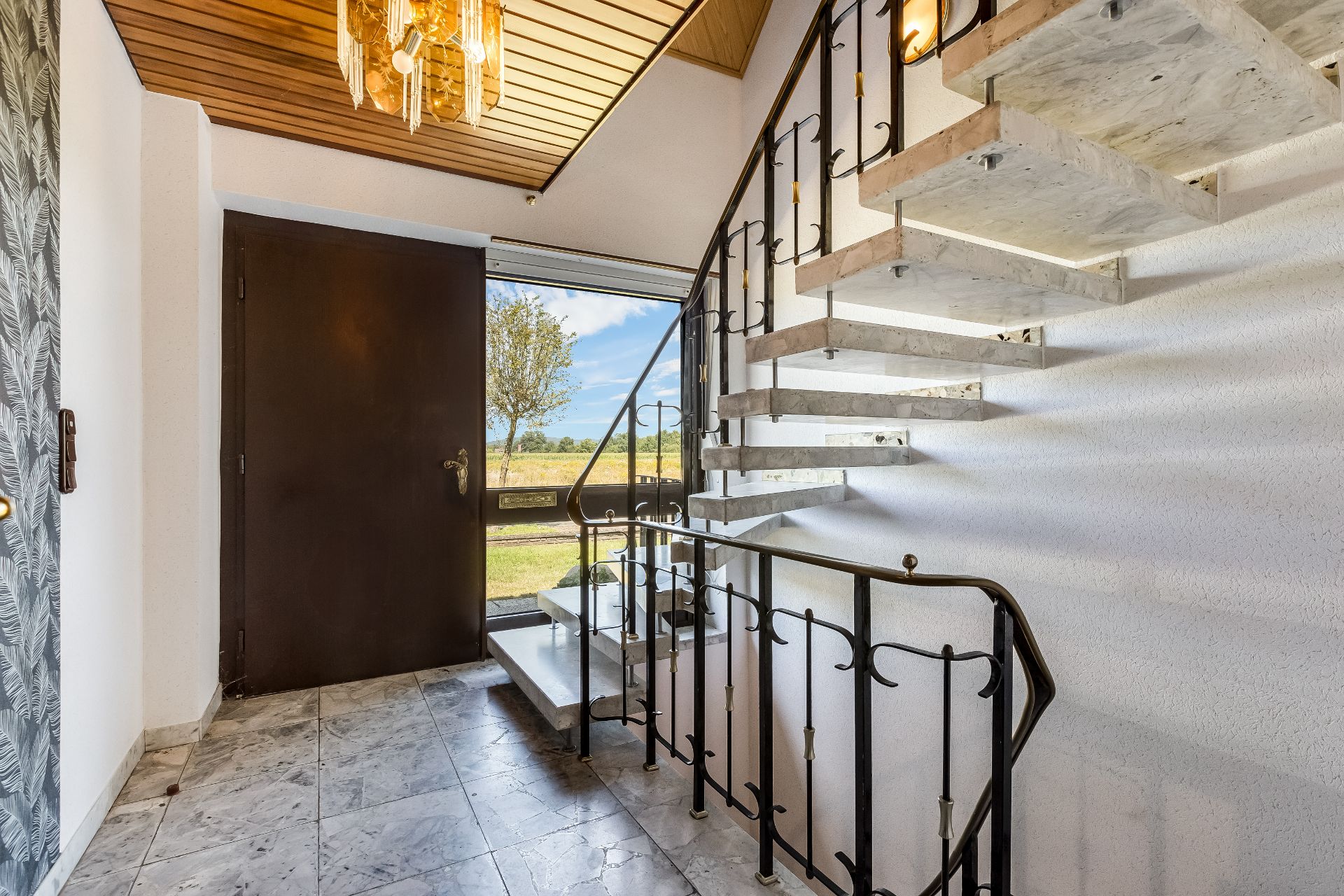 Eingangsbereich mit Marmorboden, offener Treppe und großer Fensterfront mit Blick ins Grüne.