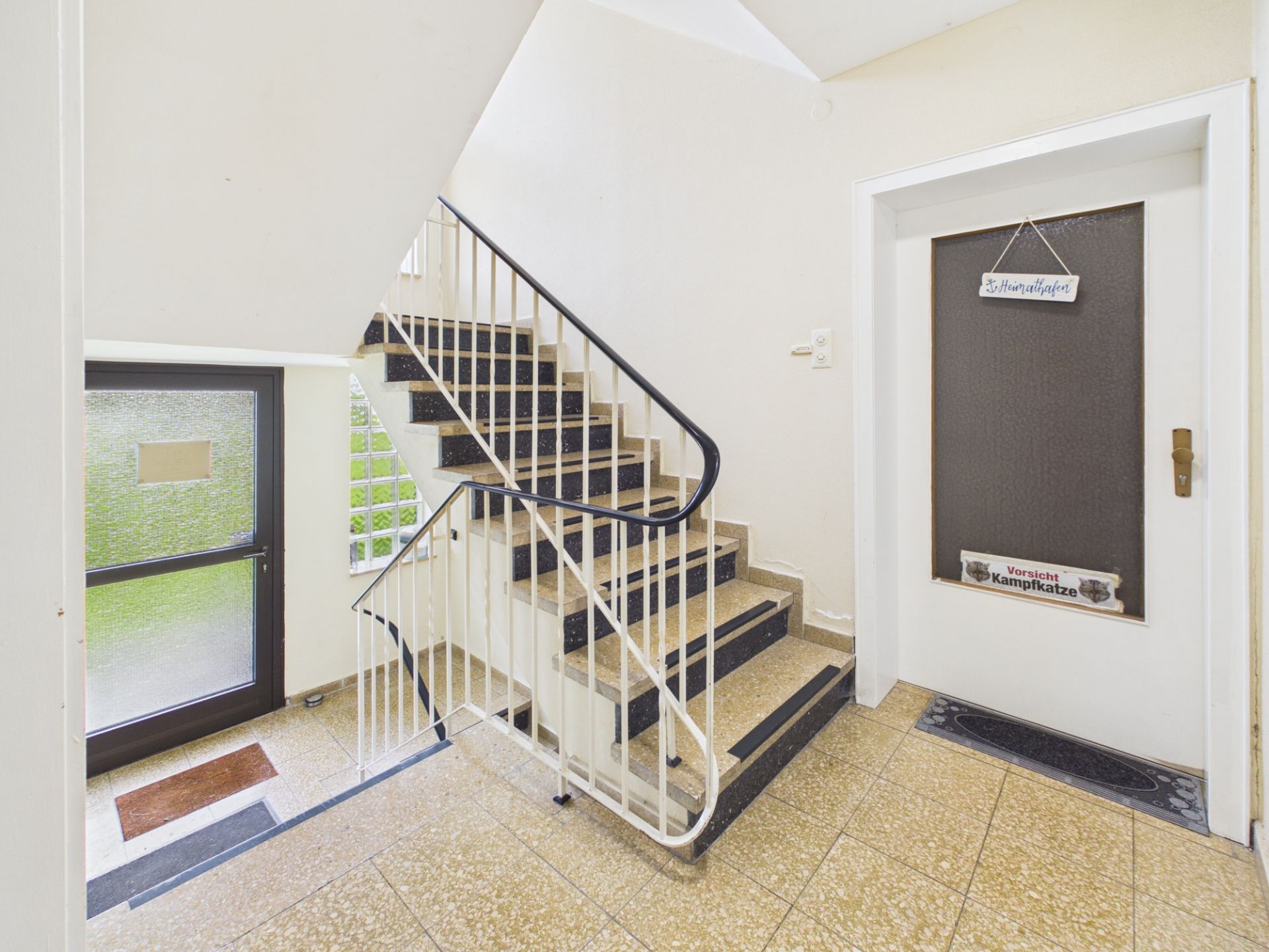 Treppenhaus mit brauner Wohnungstür, Fliesenboden und Fenstertür mit matte Verglasung.