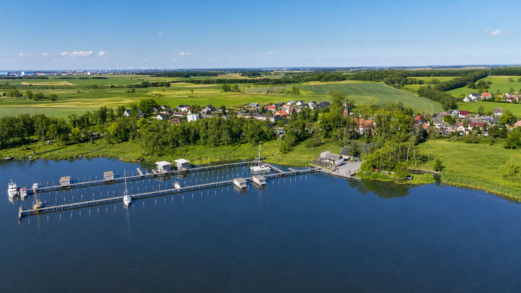 Luftaufnahme eines kleinen Hafens am See mit Booten, umgeben von Feldern und Häusern im Hintergrund.