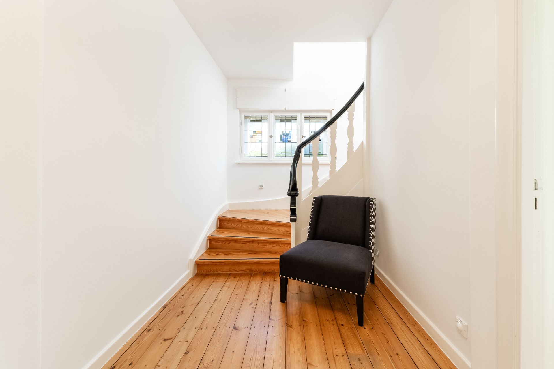 Flur mit Holzfußboden, schwarzem Stuhl und Treppe vor einem Fenster.