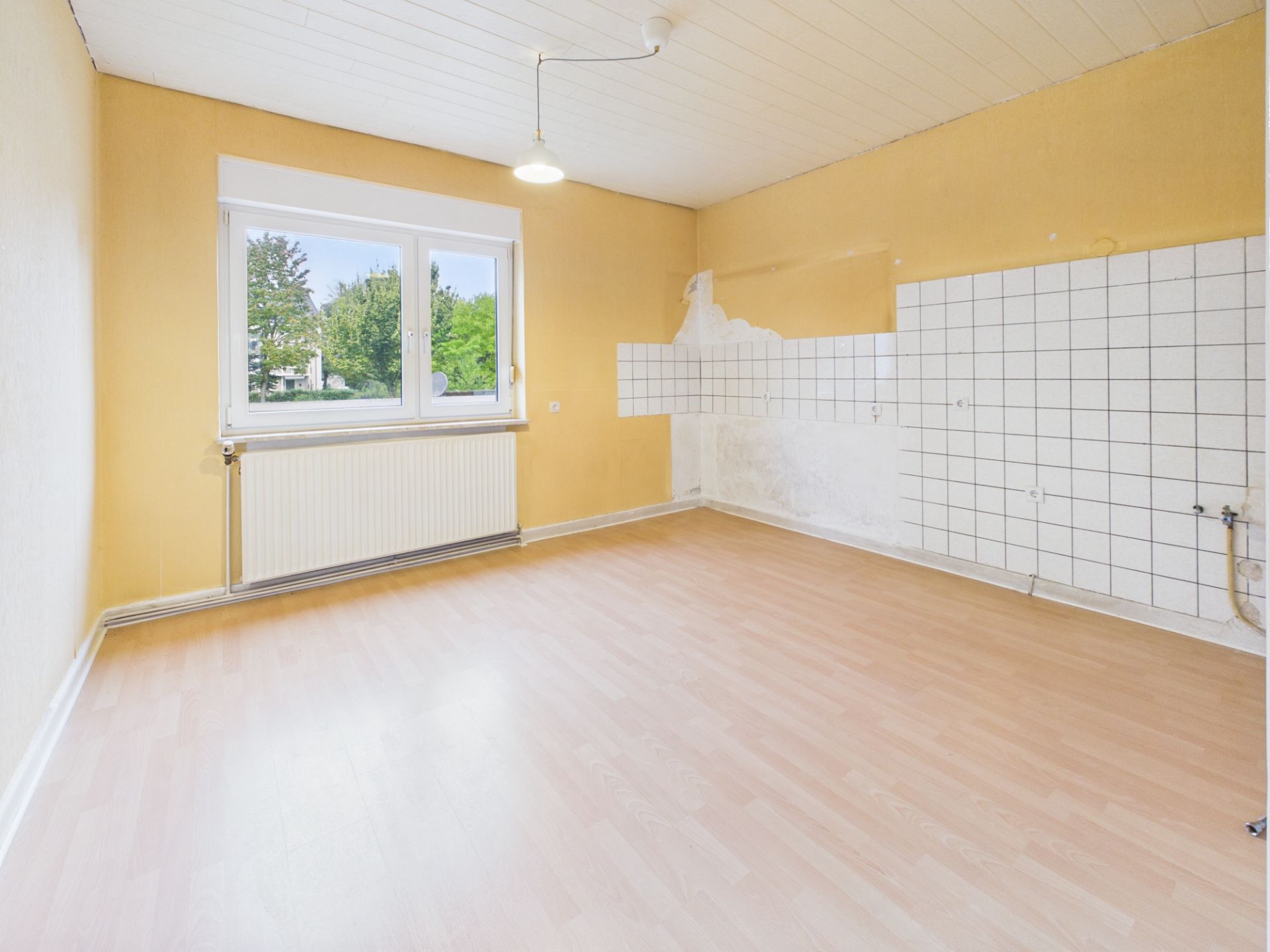 Leerer Raum mit gelben Wänden, großem Fenster, hellem Boden und weißen Wandfliesen an einer Seite.
