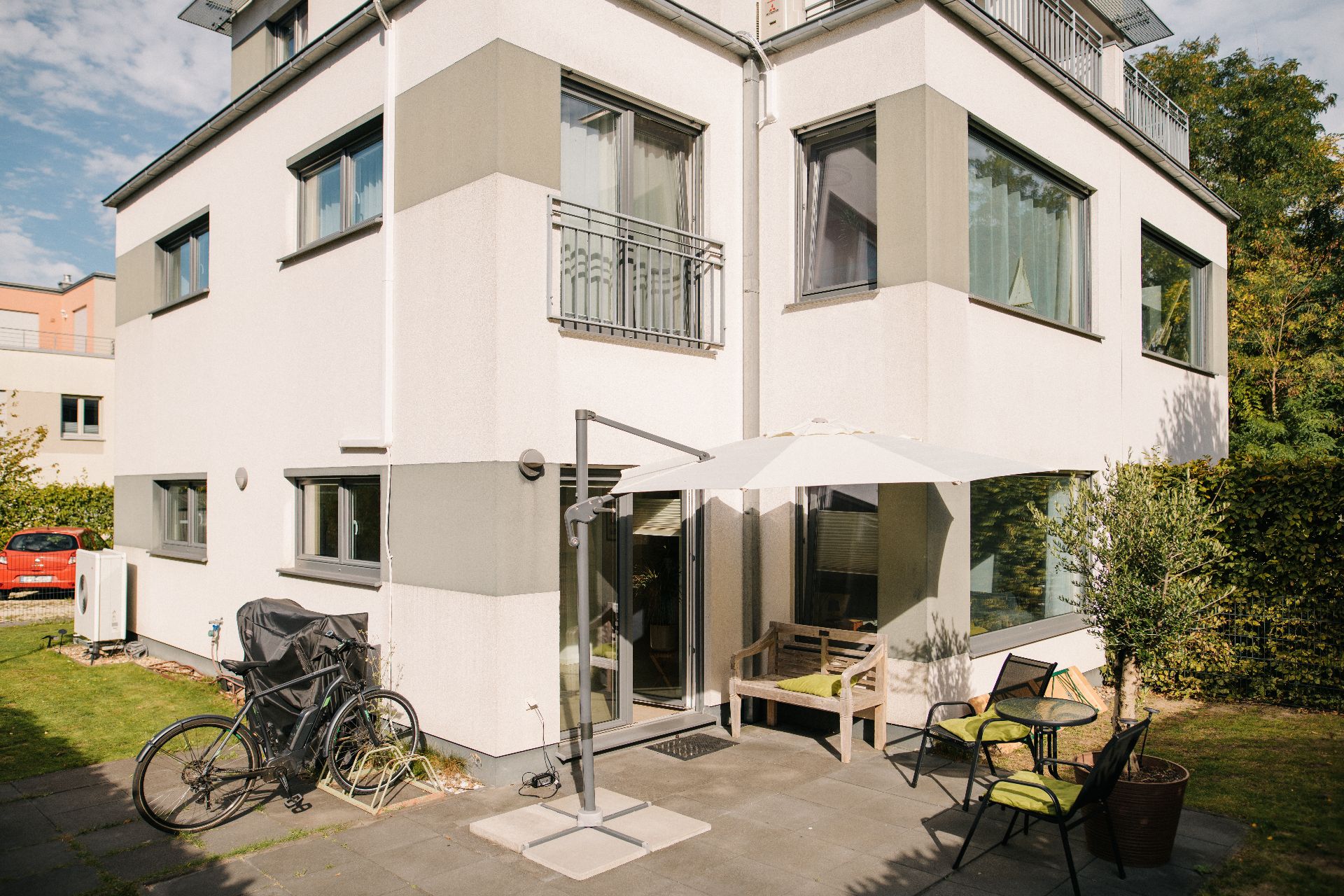 Moderne Hausfassade mit Terrasse, Sonnenschirm, Sitzbank, Stühlen und Fahrrad im Garten.