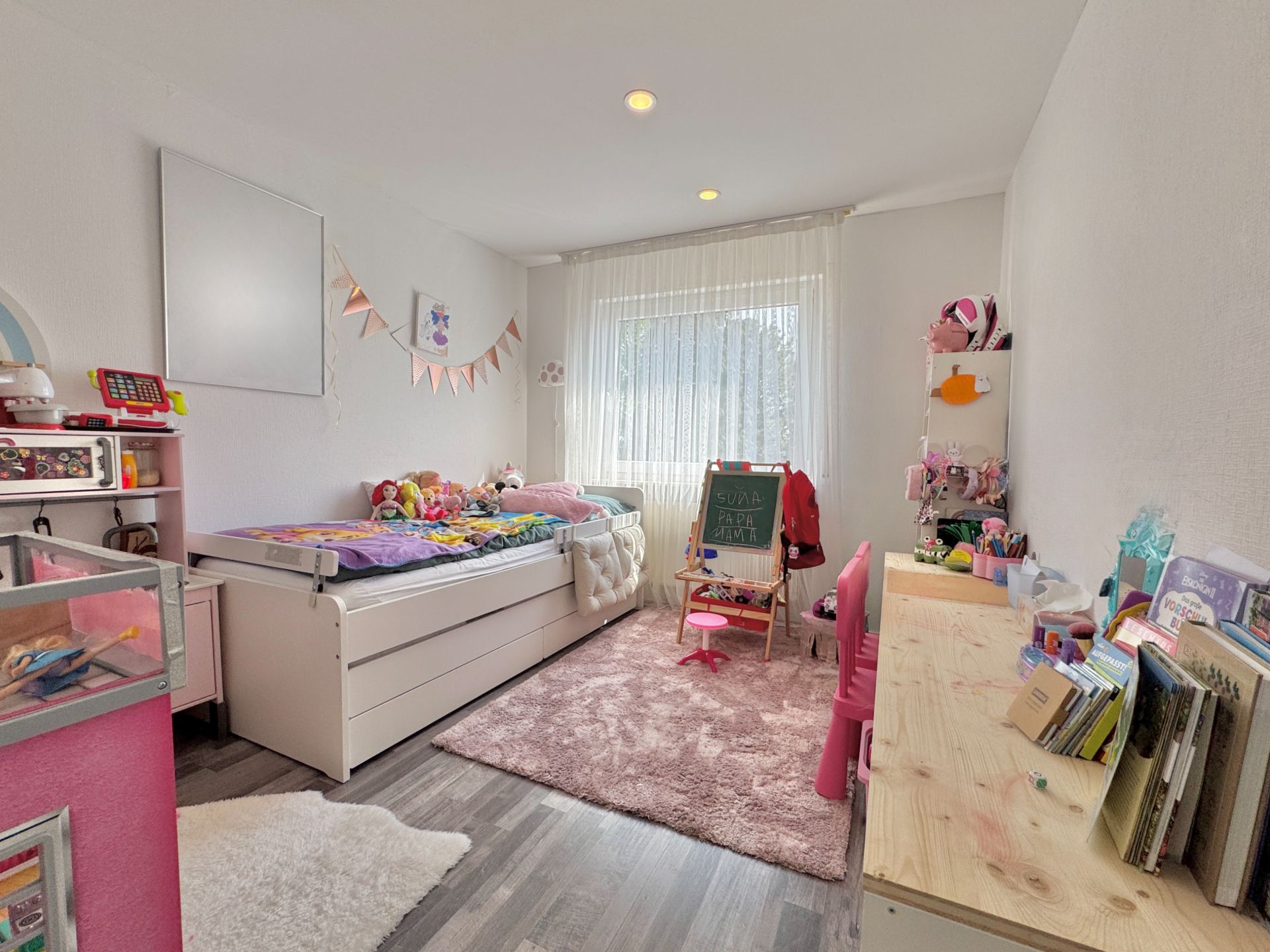 Helles Kinderzimmer mit Einzelbett, Schreibtisch, Spielzeug und großem Fenster mit Gardinen.
