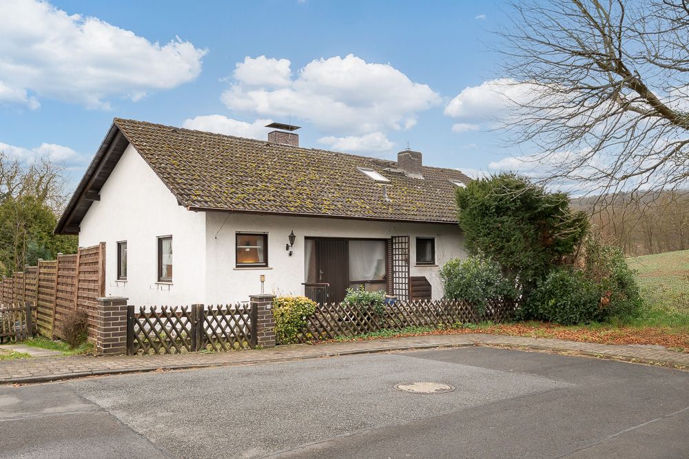 Weißes Einfamilienhaus mit Gartenzaun, Büschen und schrägem Dach an einer ruhigen Straße.