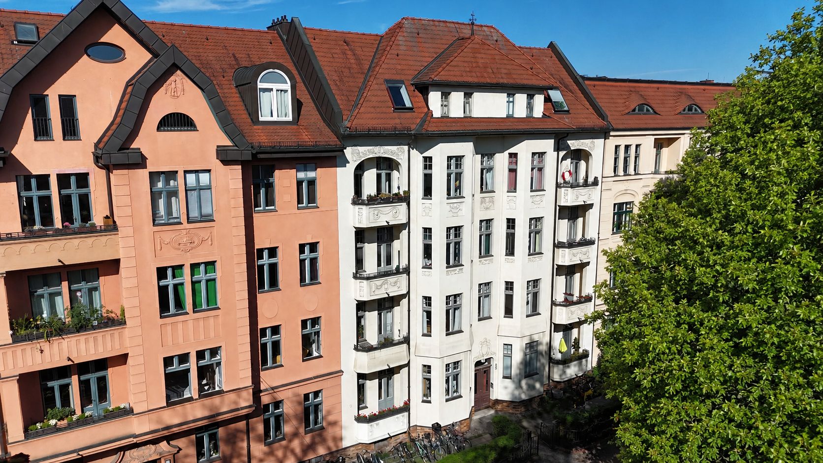 Mehrstöckiges Wohnhaus mit roten Dächern, Balkonen und vielen Fenstern, daneben ein großer Baum.