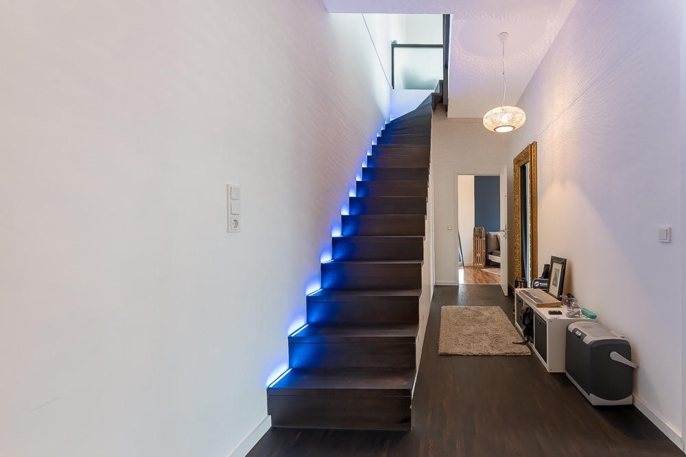 Flur mit dunkler Treppe und blauer LED-Beleuchtung, großem Spiegel und Teppich.