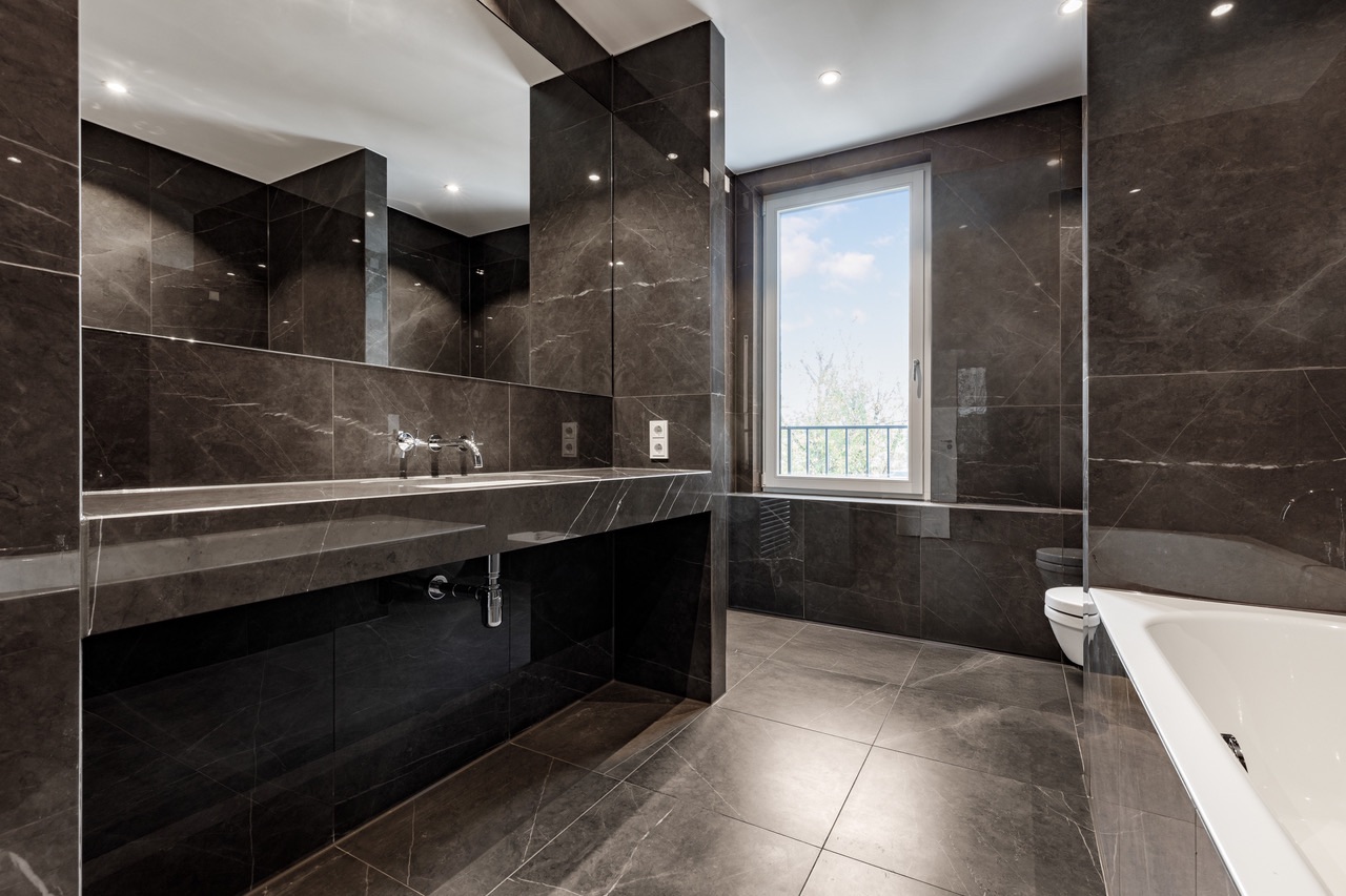 Modernes Badezimmer mit dunklen Fliesen, großem Spiegel, Waschbecken, Fenster und Badewanne.