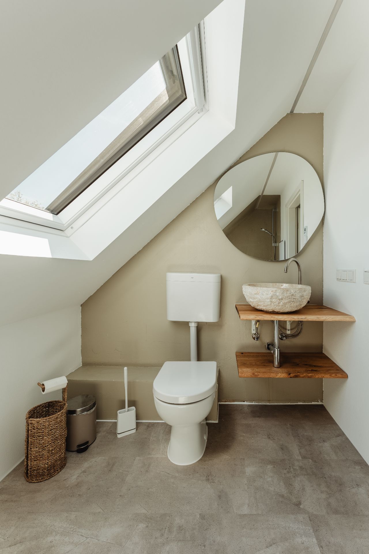 Modernes Badezimmer mit Dachfenster, Toilette, Waschbecken und rundem Spiegel.