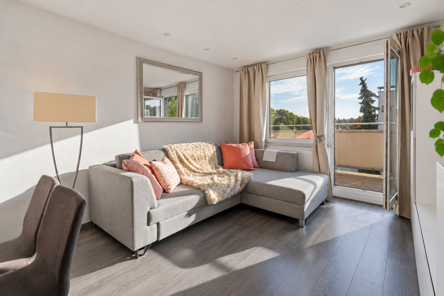 Helles Wohnzimmer mit grauem Ecksofa, Stehlampe, Spiegel und Zugang zum Balkon.