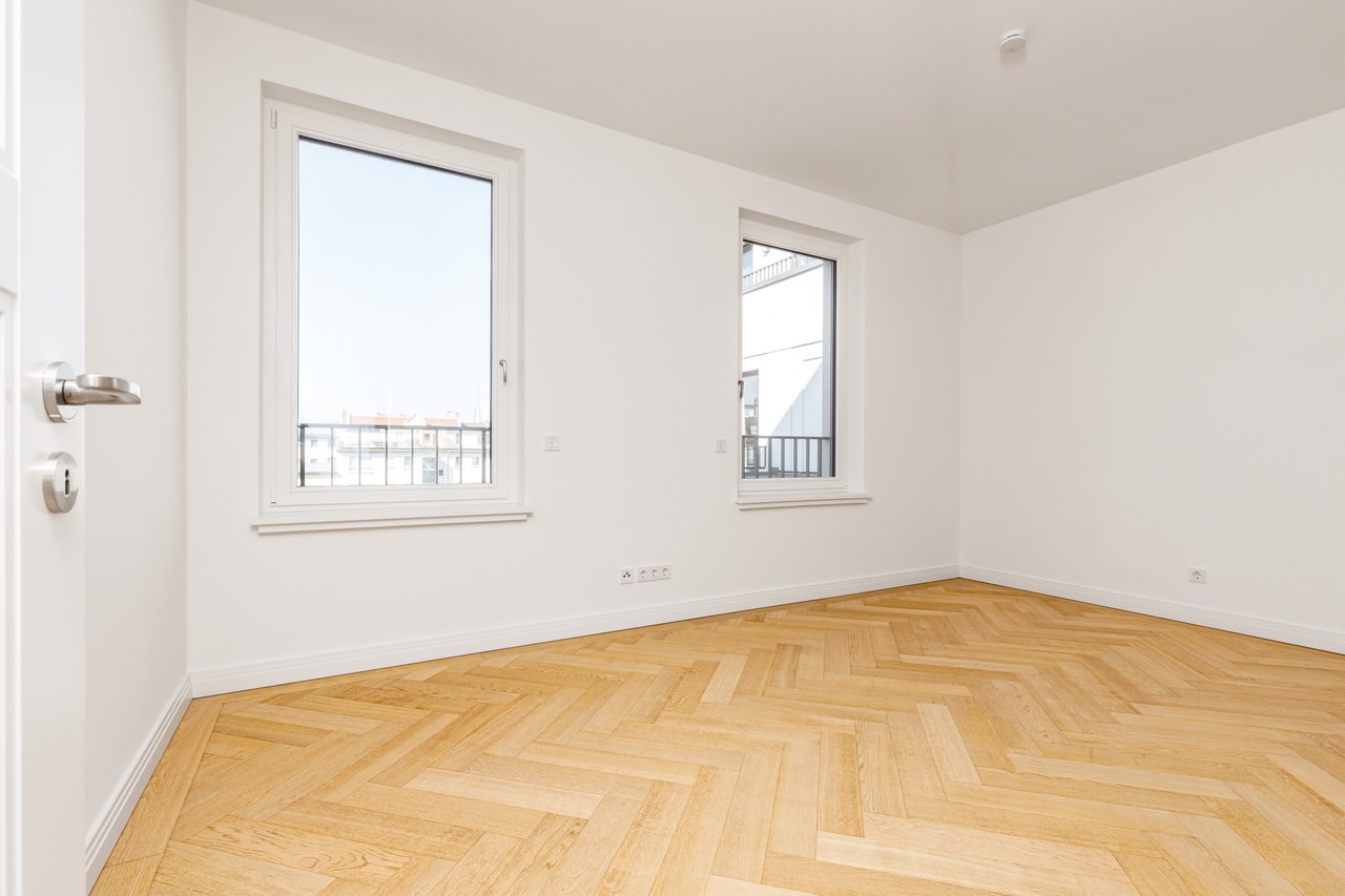 Helles, leeres Zimmer mit zwei Fenstern und Parkettboden.