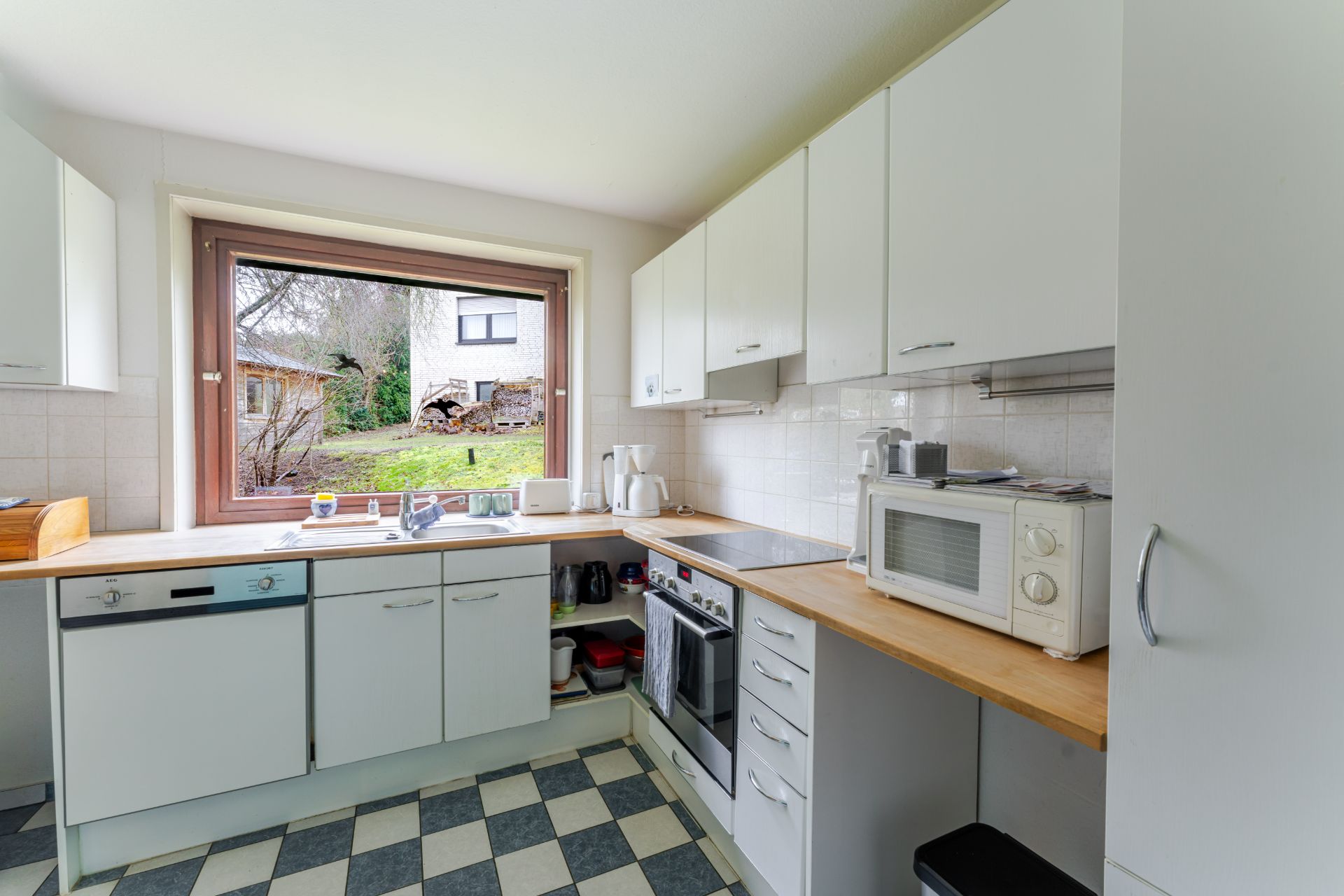 Helle Küche mit weißen Schränken, großem Fenster, Herd, Mikrowelle und Sicht auf den Garten.