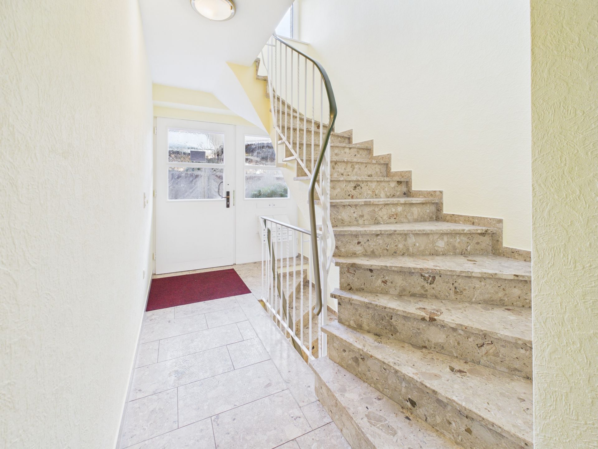 Heller Treppenhausbereich mit Steintreppe und Glastür, roter Fußmatte am Eingang.