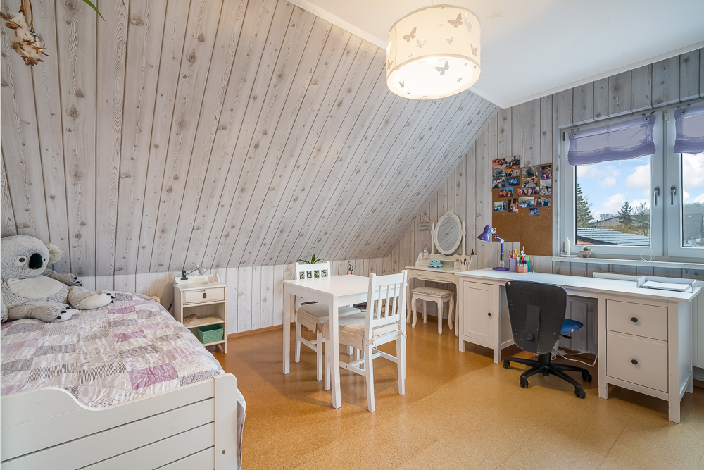 Helles Kinderzimmer mit Bett, Schreibtisch, Stuhl und Plüschtier unter schräger Holzdecke.