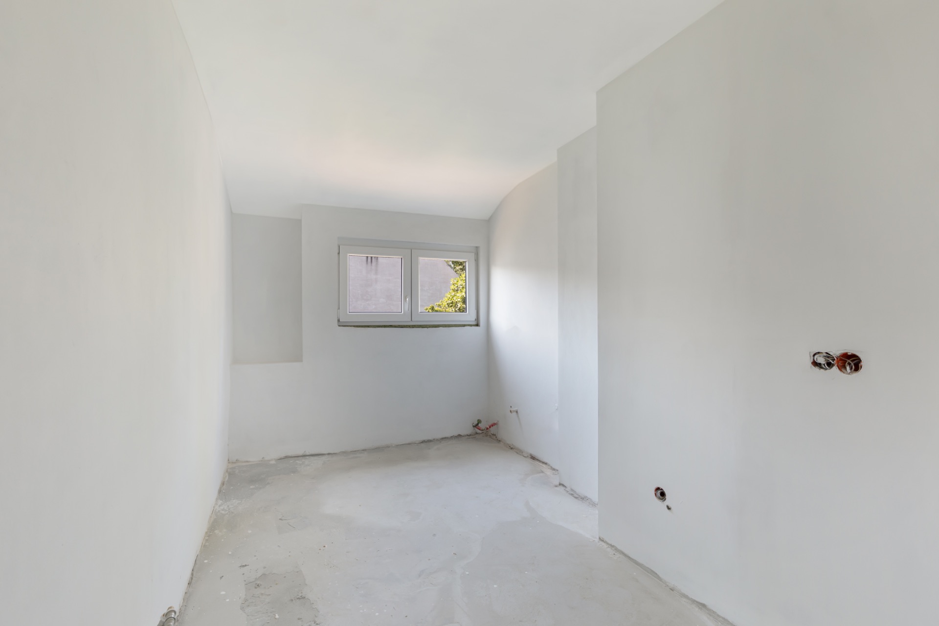 Heller, leerer Raum mit weißen Wänden, Rohboden und einem Fenster an der Stirnseite.