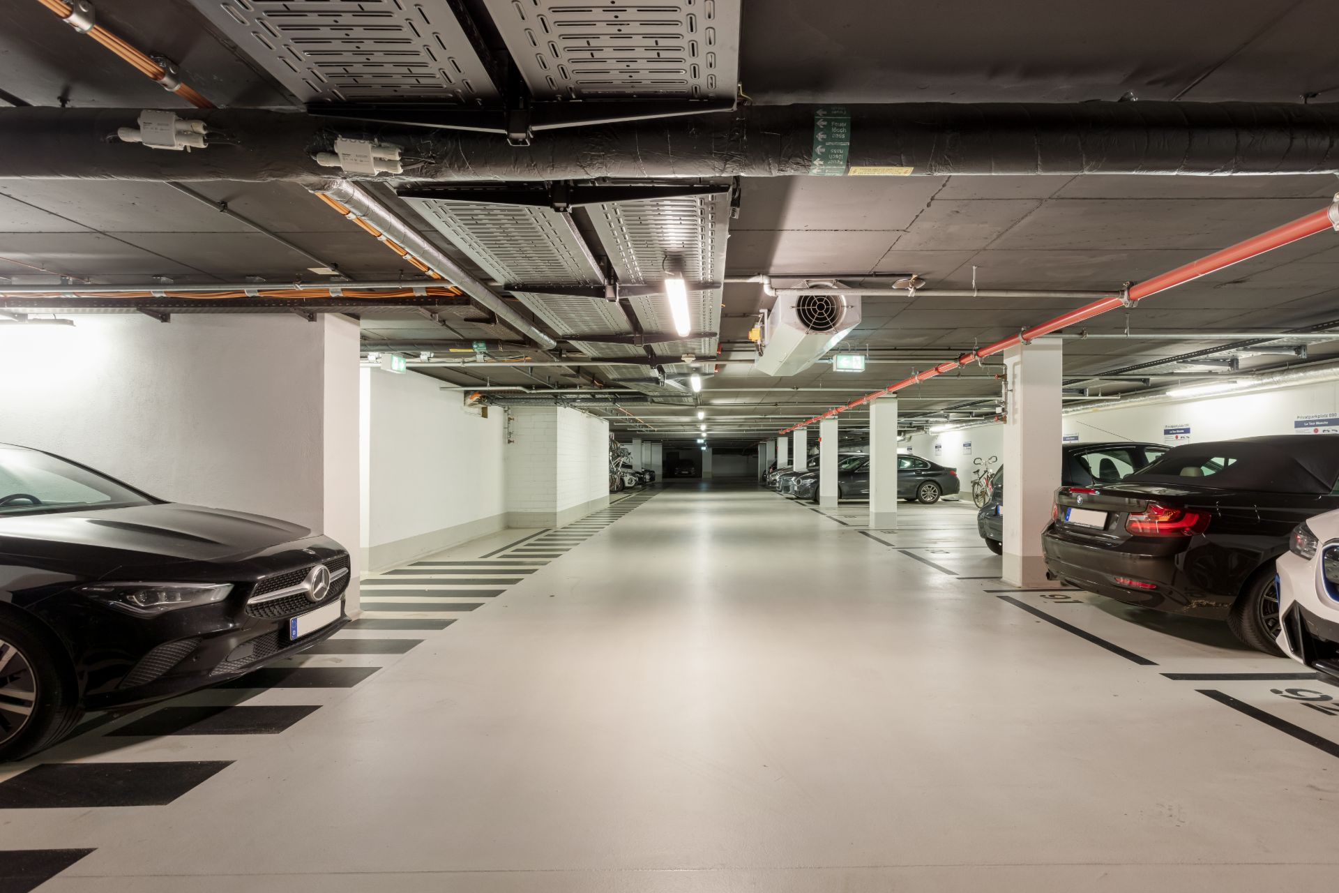 Helle Tiefgarage mit geparkten Autos und Fahrrädern entlang weißer Wände und Säulen.