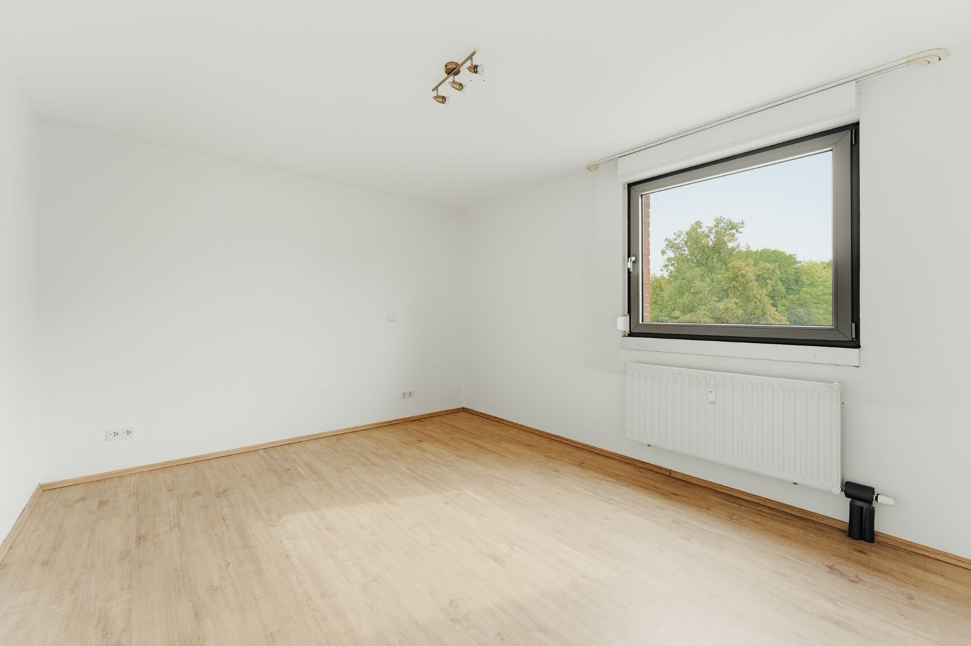 Leeres Zimmer mit hellem Holzboden, weißer Wand, Fenster und Heizkörper.