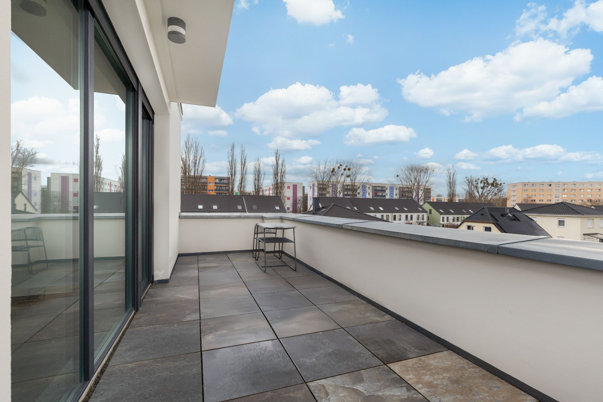 Geräumiger Balkon mit Fliesenboden, Glasgeländer und Blick auf Nachbarhäuser.