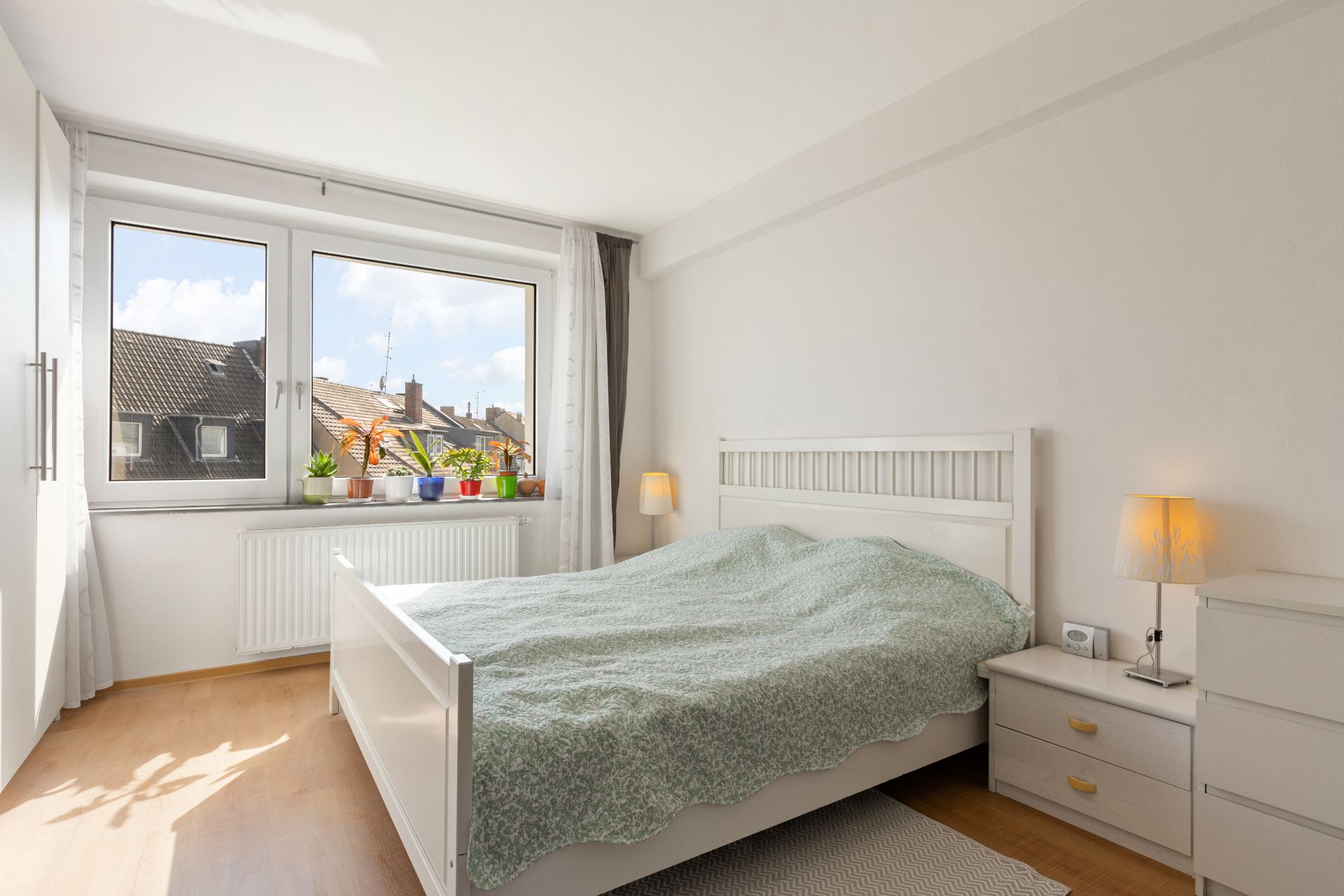 Helles Schlafzimmer mit weißem Bett, Nachttisch, Lampen und Pflanzen auf der Fensterbank.