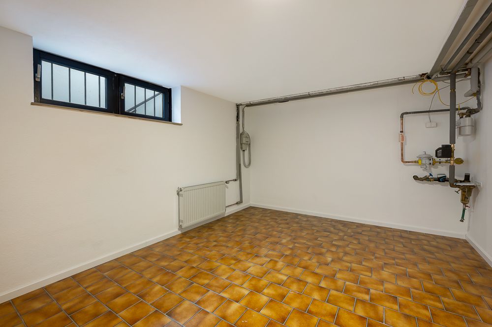 Leerer Kellerraum mit braunen Fliesen, zwei Fenstern und Heizungsrohren an der Wand.