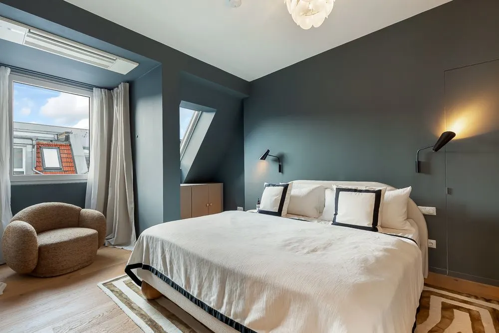 Modernes Schlafzimmer mit großem Bett, Sessel, Fenstern und dunkler Wandfarbe.