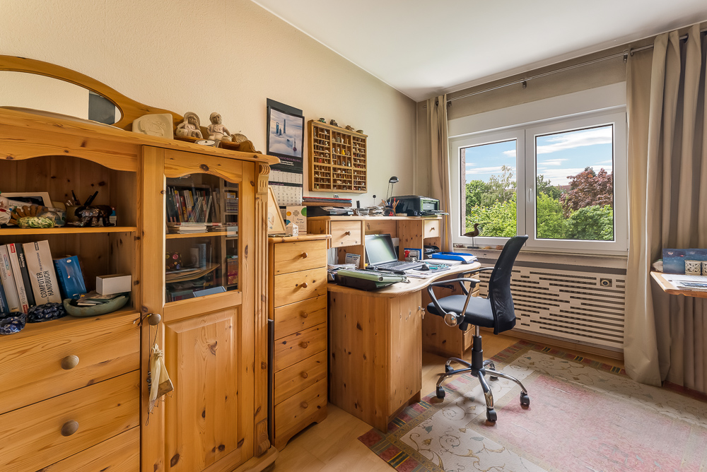 Heller Arbeitsraum mit Holzmöbeln, Schreibtisch, Regal, Stuhl und großem Fenster mit Blick ins Grüne.