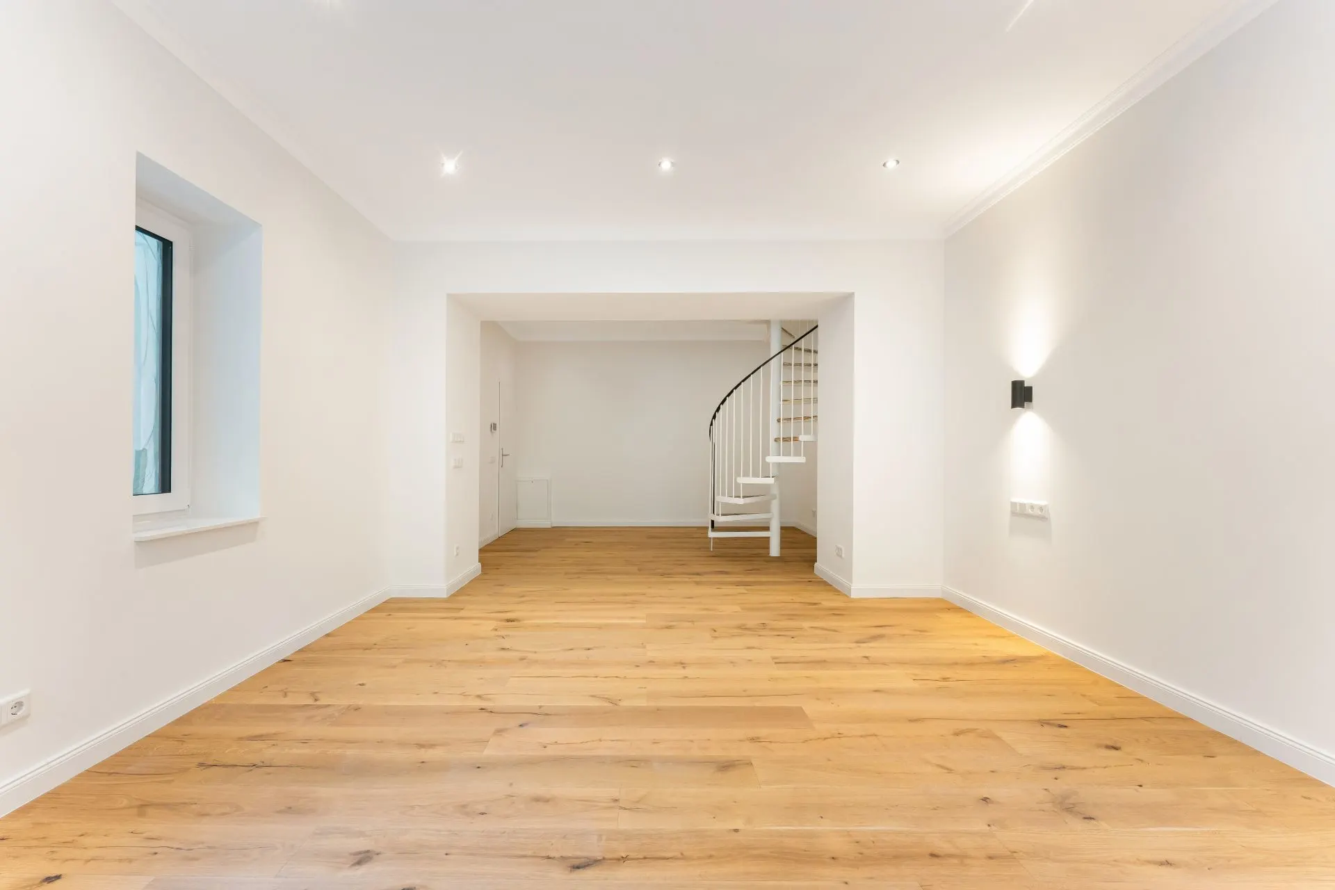 Helles Wohnzimmer mit Holzfußboden und weißer Wendeltreppe im Hintergrund.