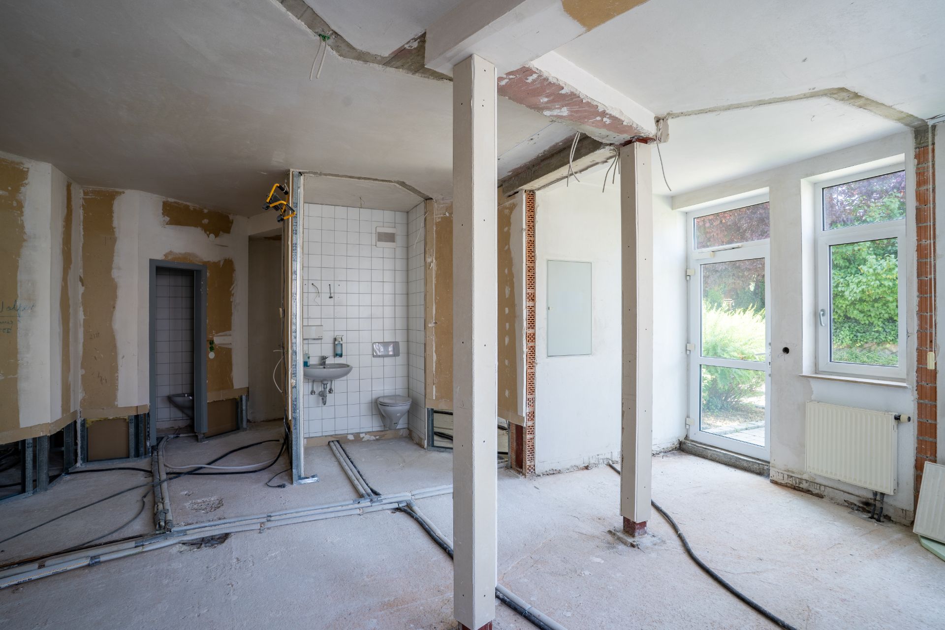 Renovierungsbedürftiger Raum mit sichtbaren Rohren, Badezimmer und Ausgang zum Garten.