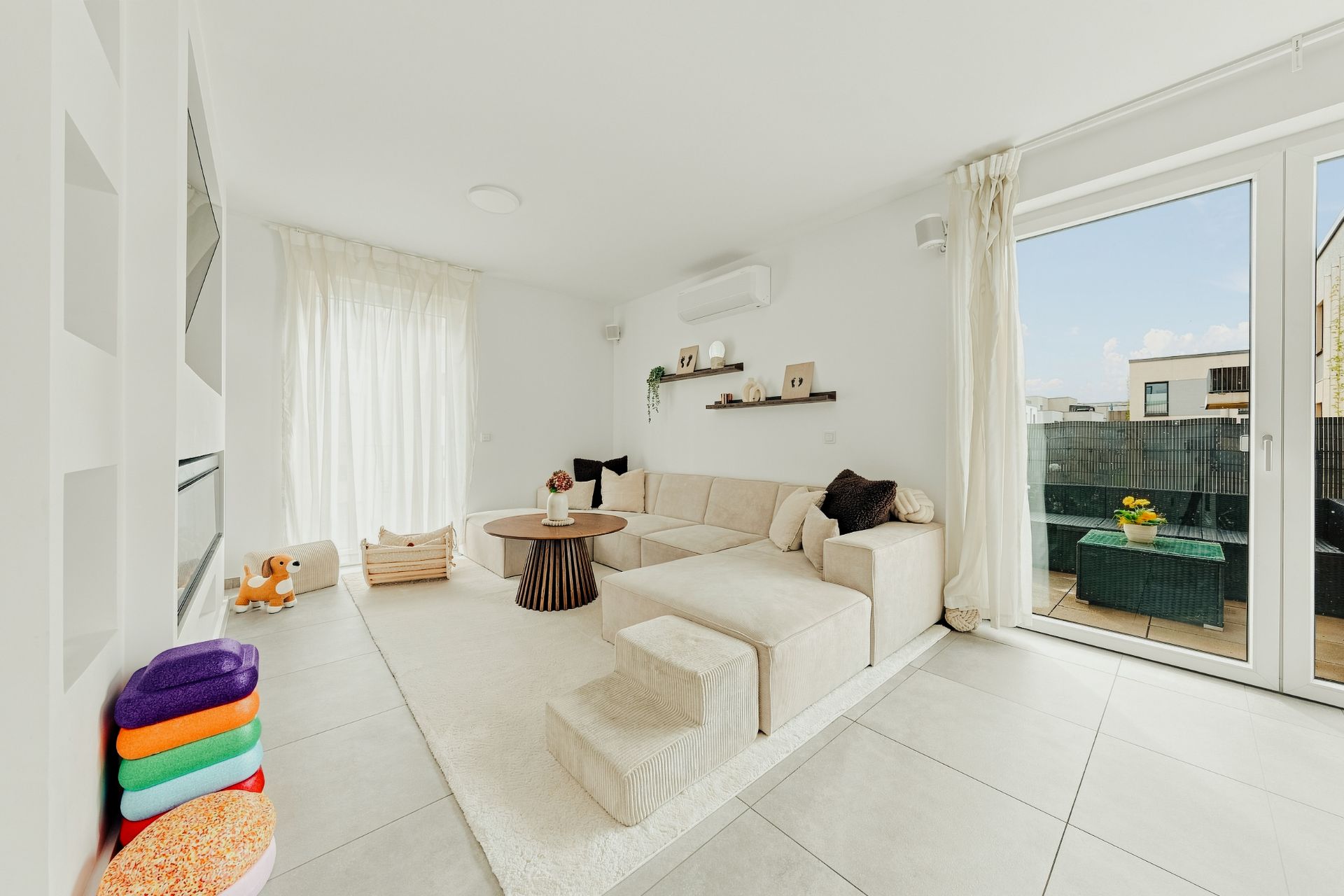 Helles Wohnzimmer mit Ecksofa, Couchtisch, Spielzeug und Zugang zum Balkon durch große Glastüren.