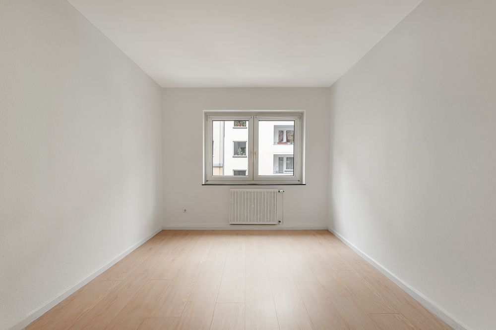 Leerer Raum mit hellem Holzboden, weißer Wand und Fenster mit Heizkörper darunter.