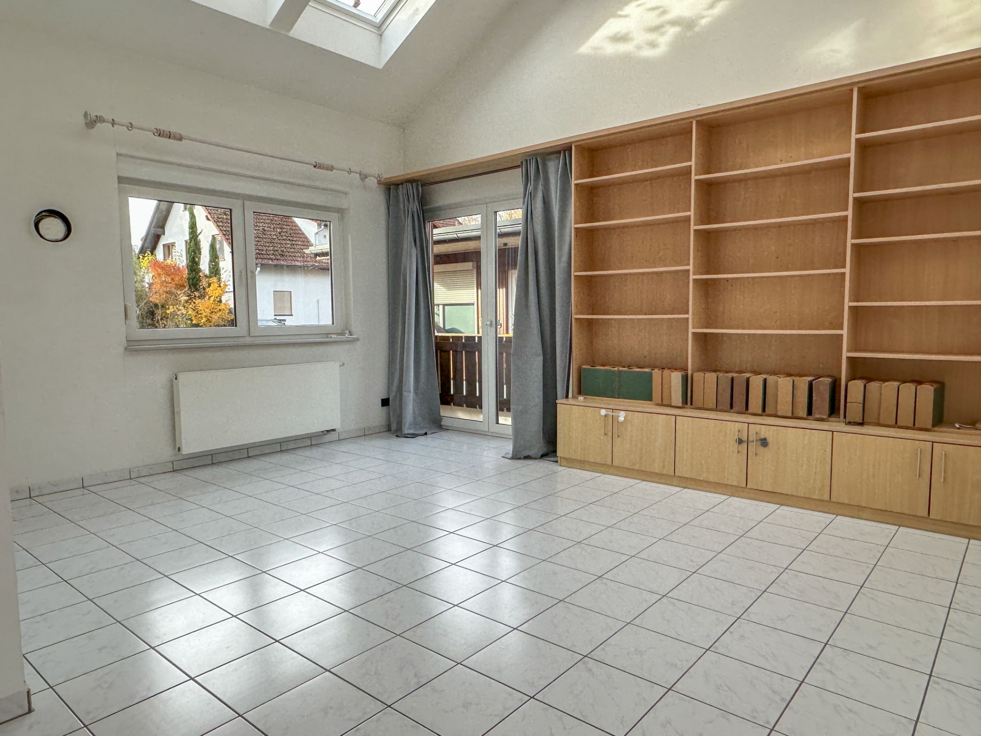 Helles Wohnzimmer mit Fliesenboden, Regalwand, Fenster und Tür zum Balkon.