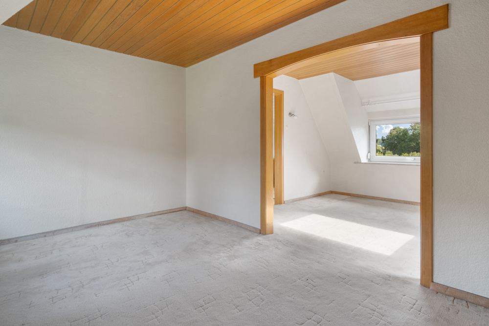 Helles, leeres Zimmer mit Teppichboden und Holzdecke. Offener Durchgang führt zu weiterem Raum mit Fenster.