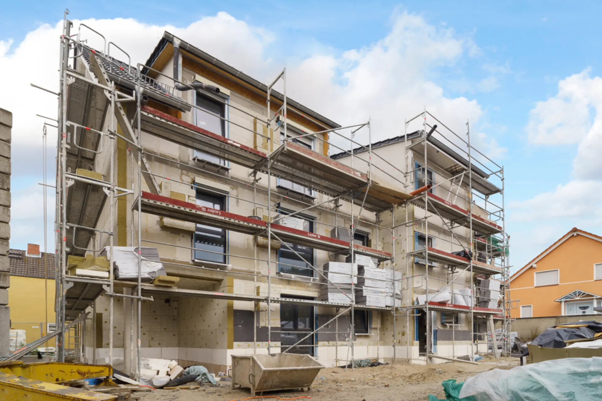 Mehrfamilienhaus im Bau mit Gerüst an der Fassade, Baumaterialien im Vordergrund.