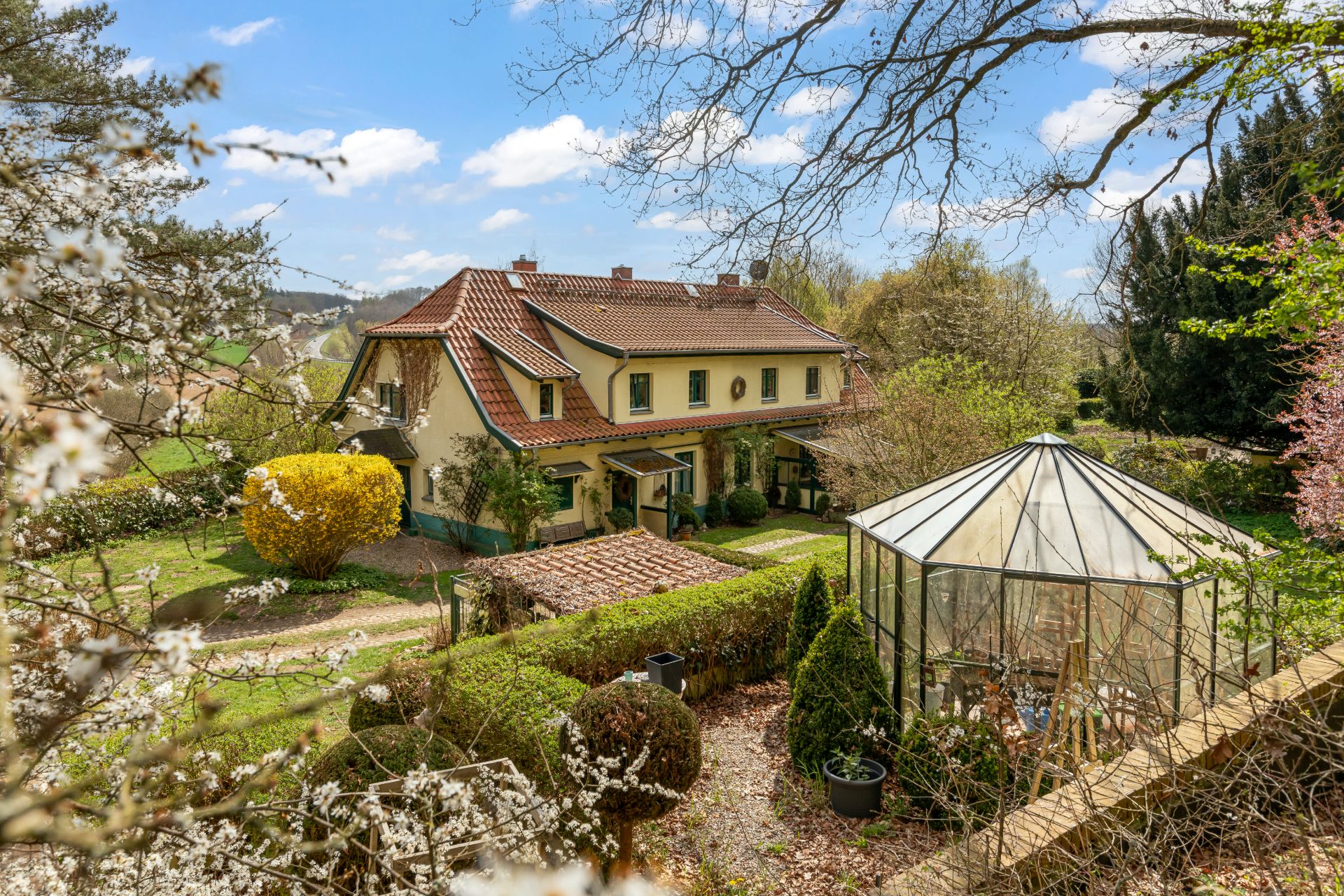 Großes Haus mit rotem Dach, Garten und Gewächshaus, umgeben von Bäumen und blühenden Sträuchern.