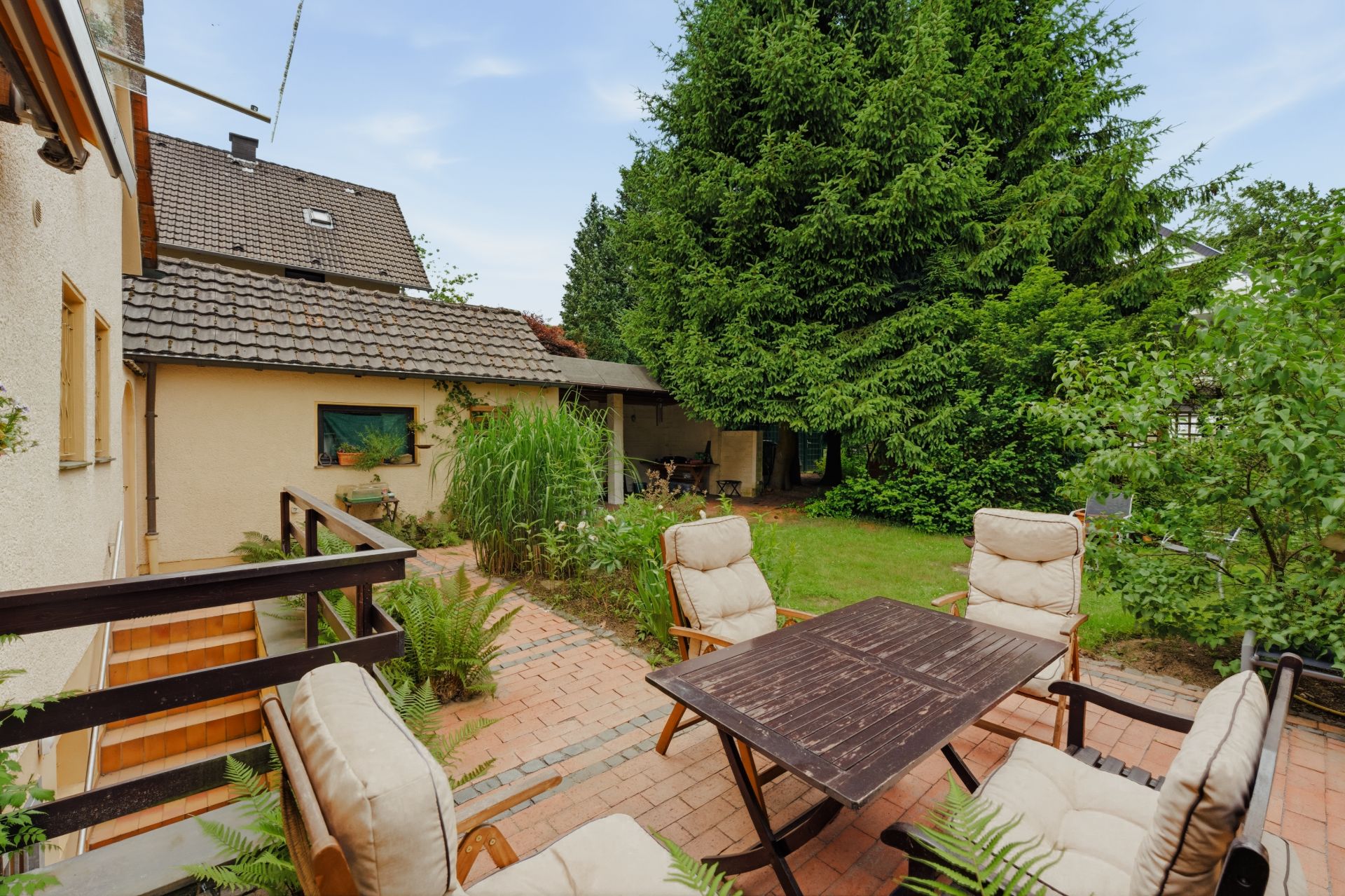 Garten mit Terrasse, Holztisch und vier gepolsterten Stühlen, umgeben von Bäumen und Pflanzen.