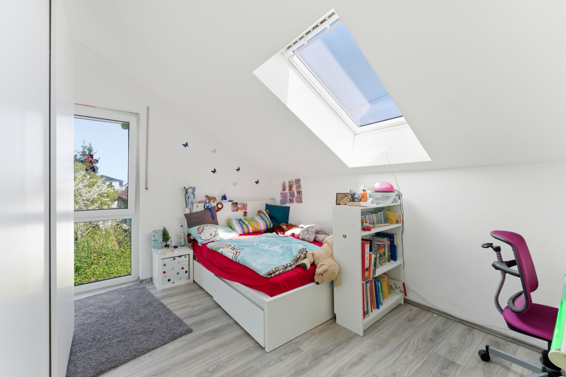 Helles Kinderzimmer mit Einzelbett, Dachfenster, Bücherregal und Schreibtisch unter einer Schräge.