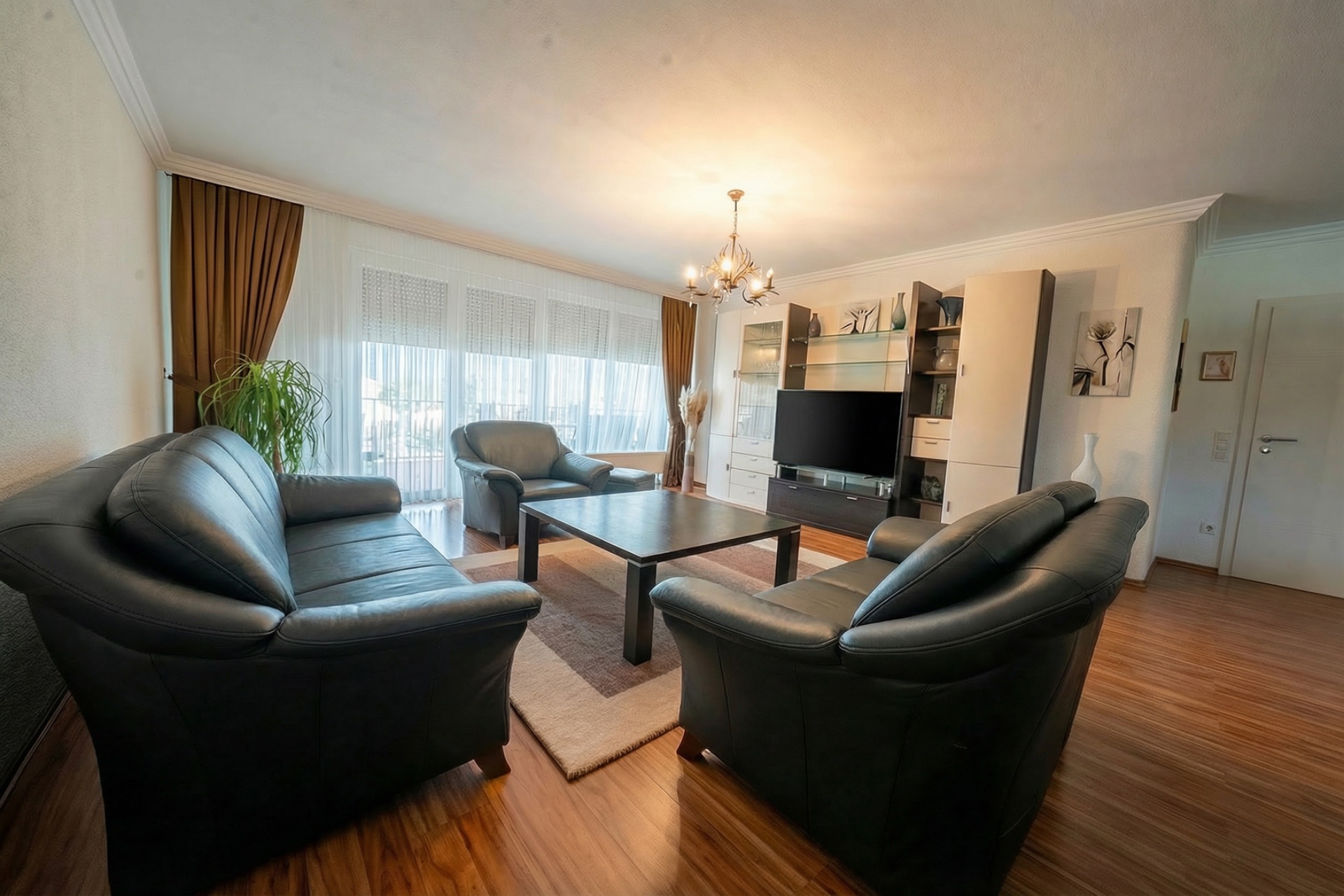 Wohnzimmer mit drei Ledersofas, Couchtisch, TV und großen Fenstern mit Vorhängen.