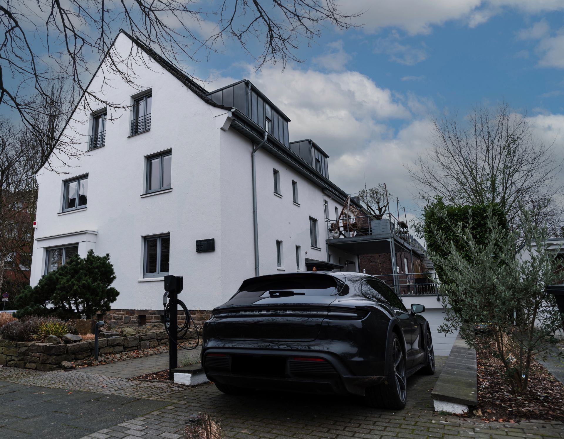 Weißes Wohnhaus mit grauem Dach, schwarzem Auto und Ladestation im Vorgarten.