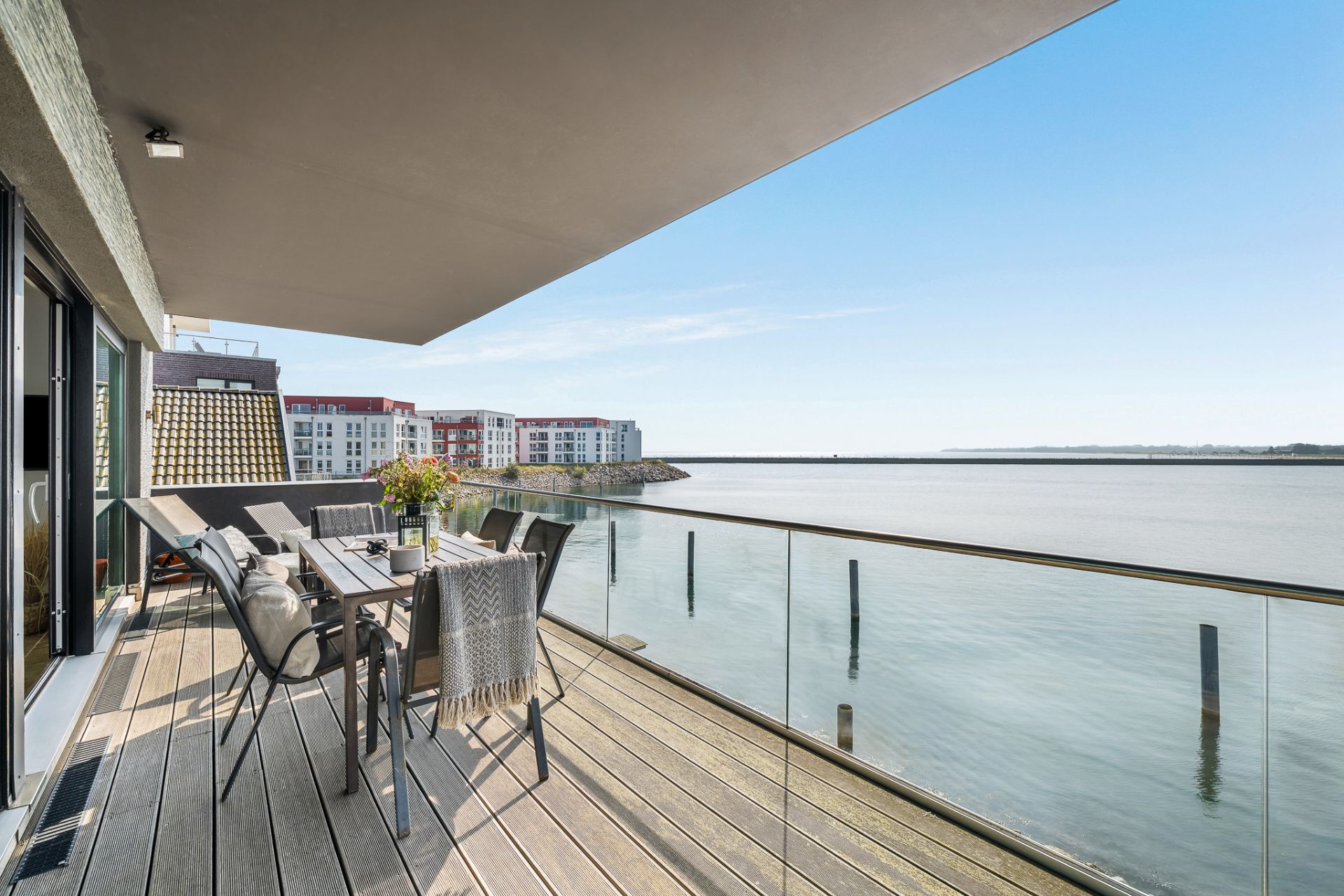 Großer Balkon mit Esstisch und Stühlen, Blick auf das Wasser und moderne Wohngebäude im Hintergrund.