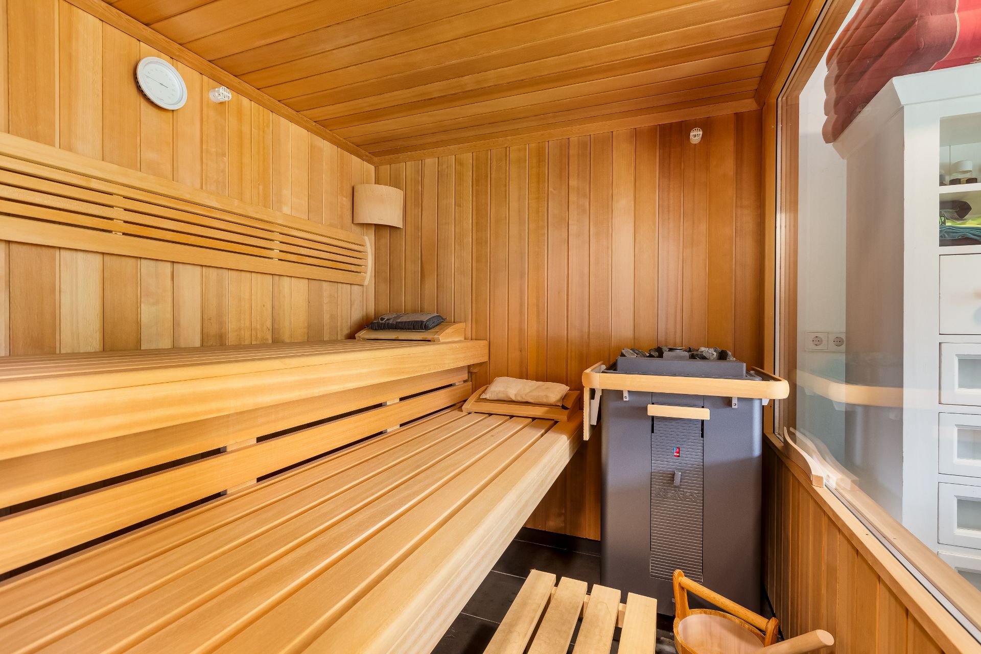 Holzsauna mit Bänken, Saunaofen, Handtüchern und großem Fenster zu einem anderen Raum.