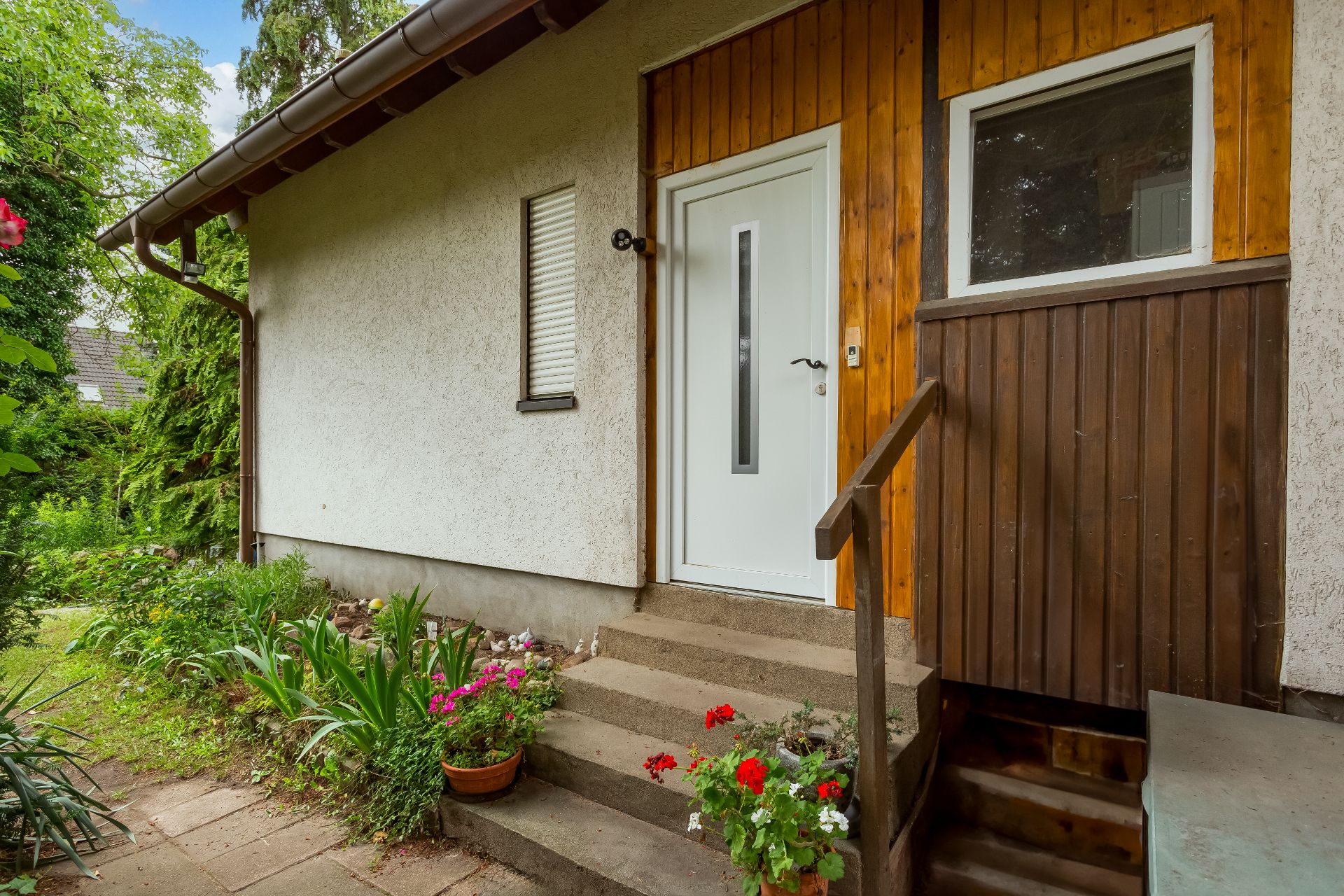 Eingangstür eines Hauses mit Treppe, Blumenbeeten und Blumentöpfen im Vorgarten.