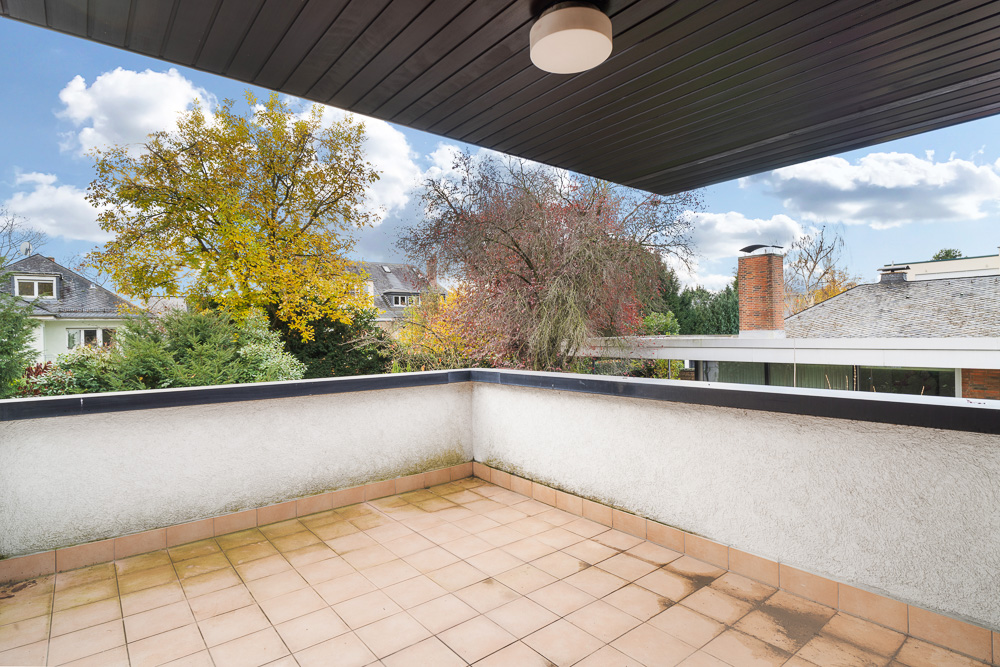 Große, überdachte Terrasse mit Fliesenboden und Blick auf Bäume und Nachbarhäuser.